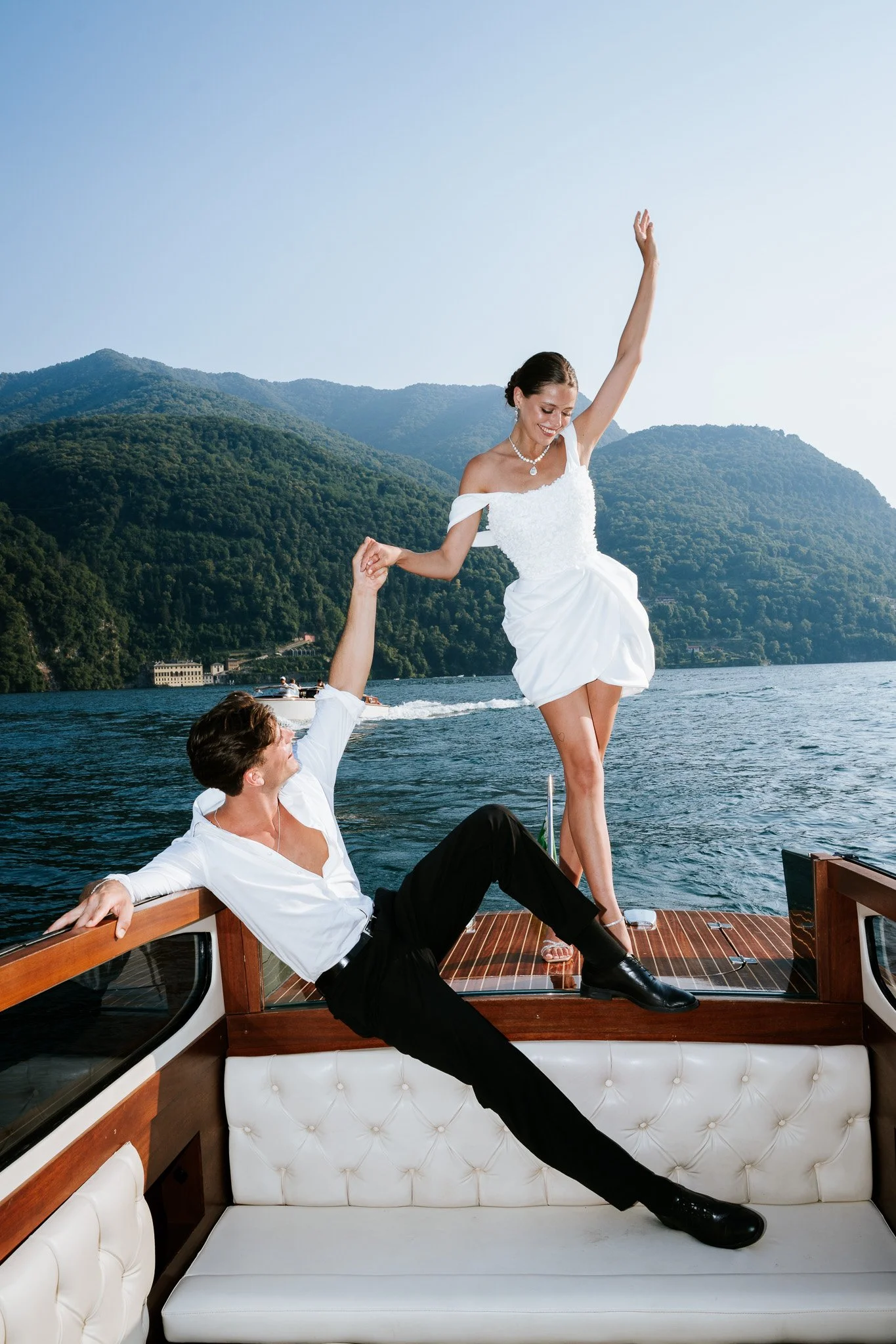A woman in a white dress and pearl necklace standing on a boat with water, mountains, and a boat in the background, while holding hands with a man sitting on the boat with his back against the railing.