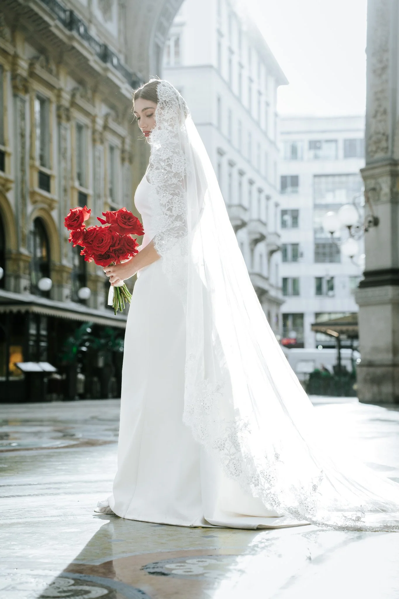 A bride in a white wedding dress holding a bouquet of red roses, standing outdoors in a city with historic buildings in the background.