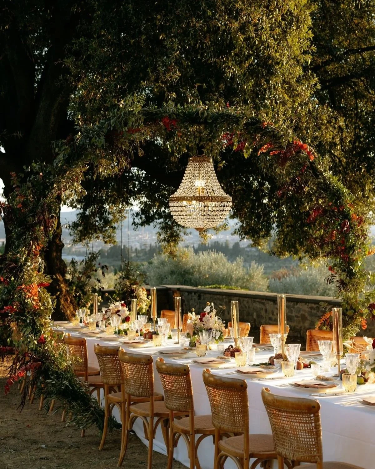 An outdoor dining table set under a large tree with a chandelier hanging from its branches, decorated with a floral arch, and a scenic landscape in the background.