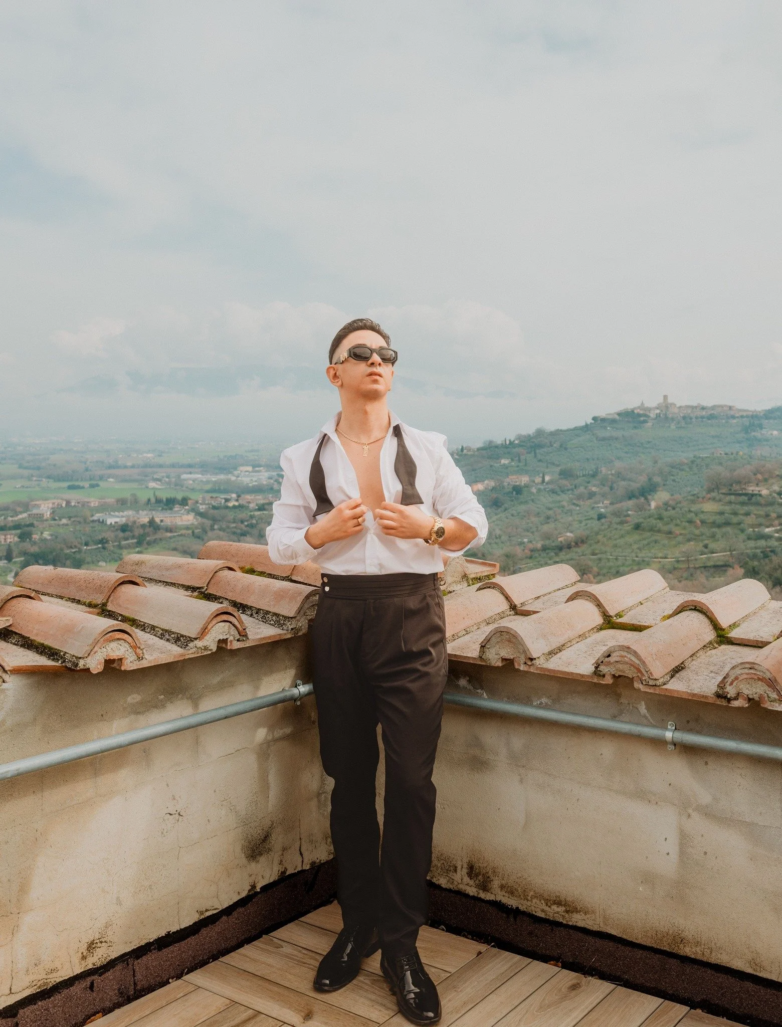 Young person standing on a rooftop with a city and hilly landscape in the background, wearing sunglasses, black pants, a white shirt, and a loosened black tie.