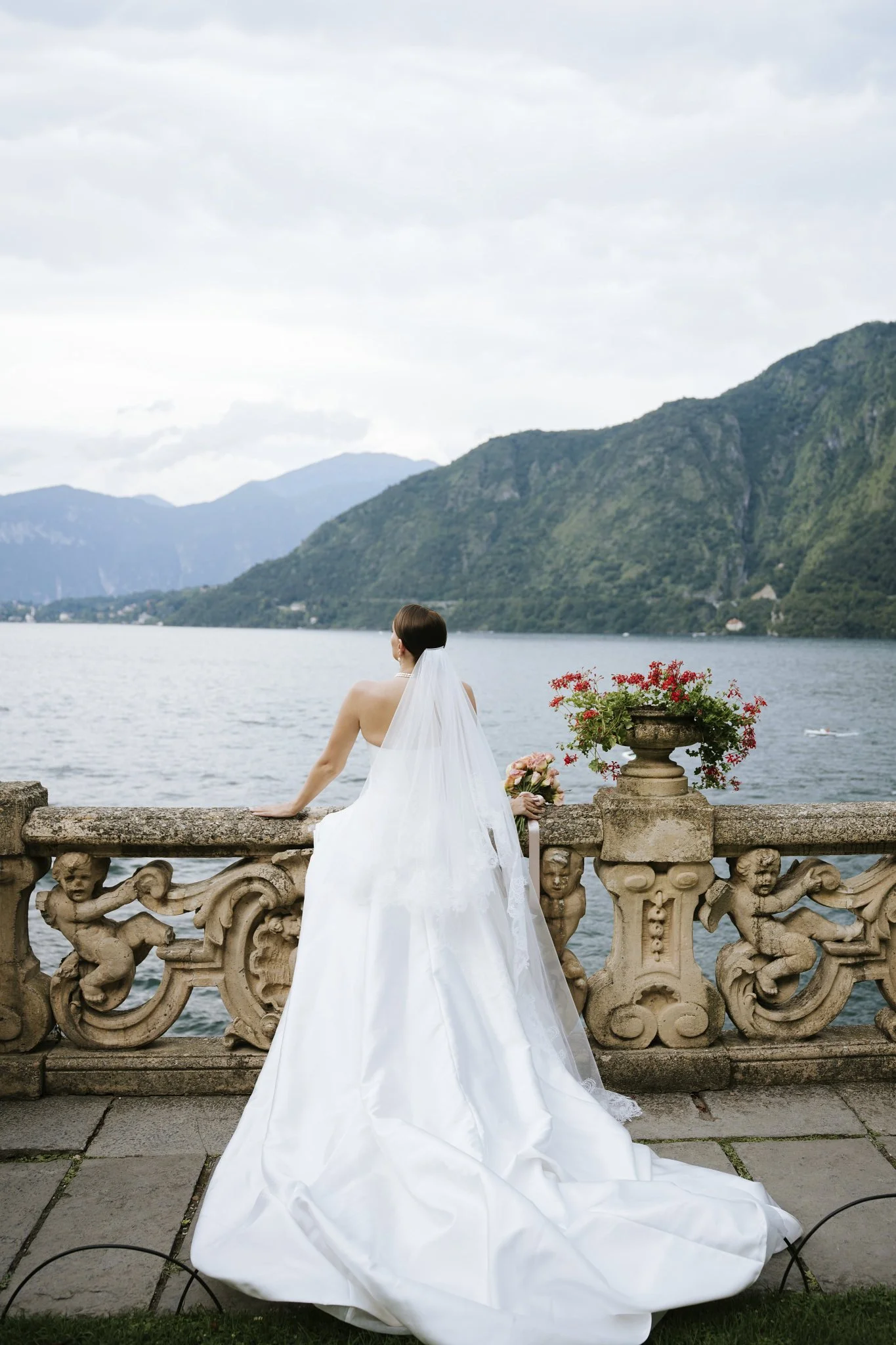 Bride sitting on the stone balustrade at Villa del Balbianello, Lake Como, with the water and mountains in the background.