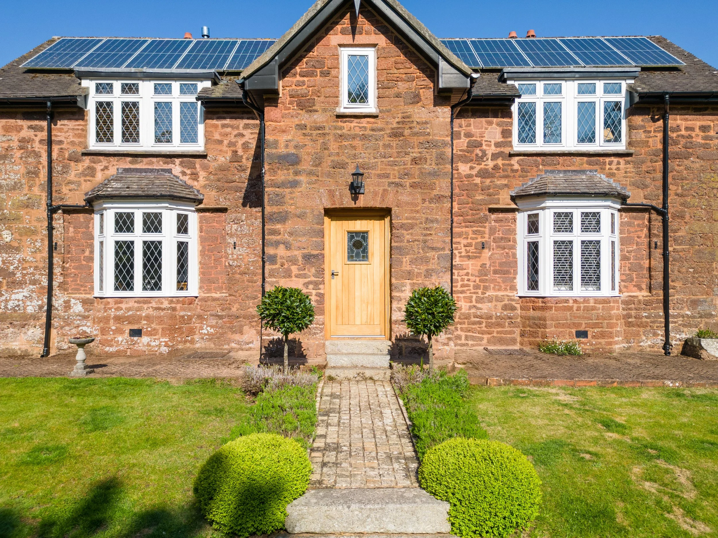 A brick house with white-framed bay and single windows, black downspouts, a wooden front door, and a front yard with a stone pathway, trimmed bushes, a lawn, and a clear blue sky.