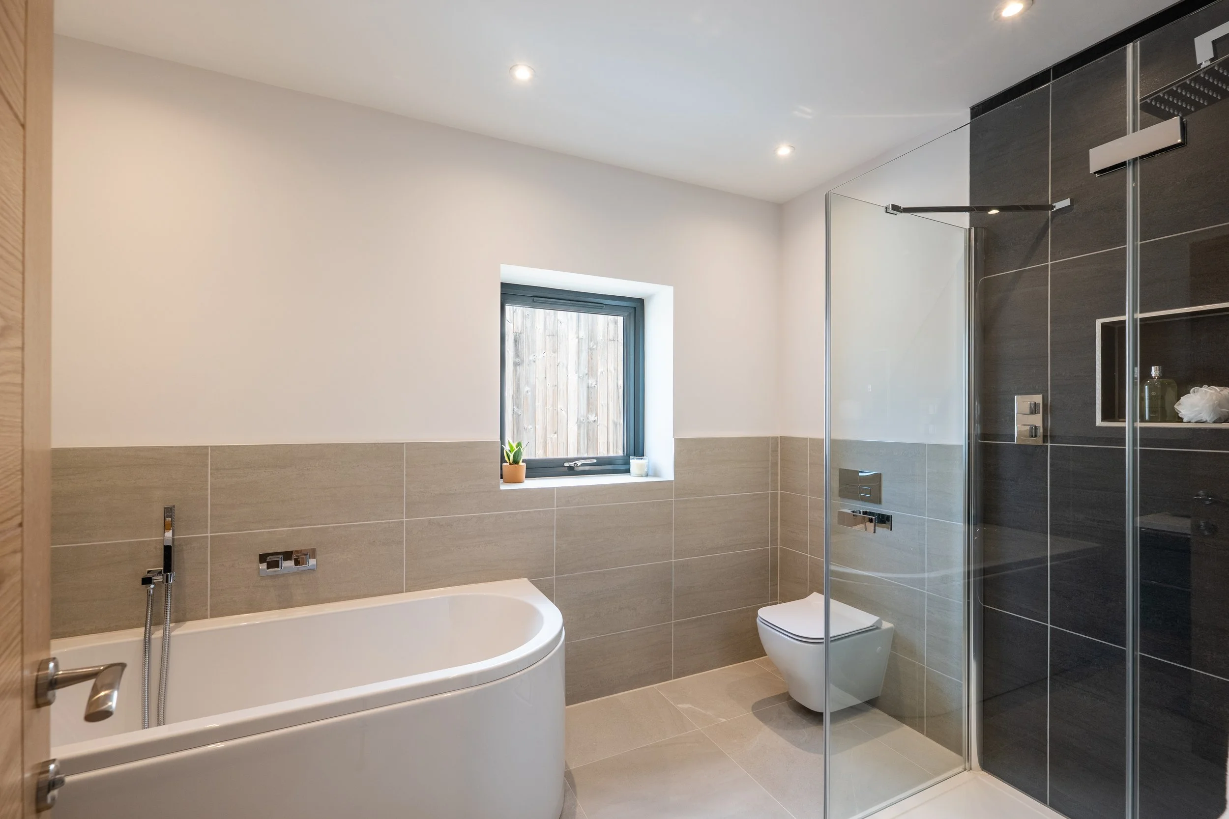 Modern bathroom with a white bathtub, a frosted window with a small potted plant and candle, a white toilet, and a walk-in shower with dark tiles and glass door.