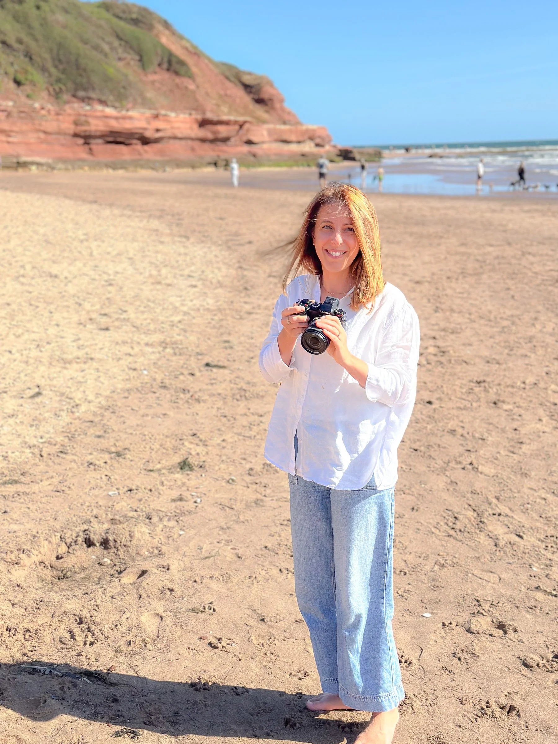 A woman with light brown hair smiling and holding a camera on a sandy beach with cliffs in the background. Other people are walking along the shore and enjoying the day.