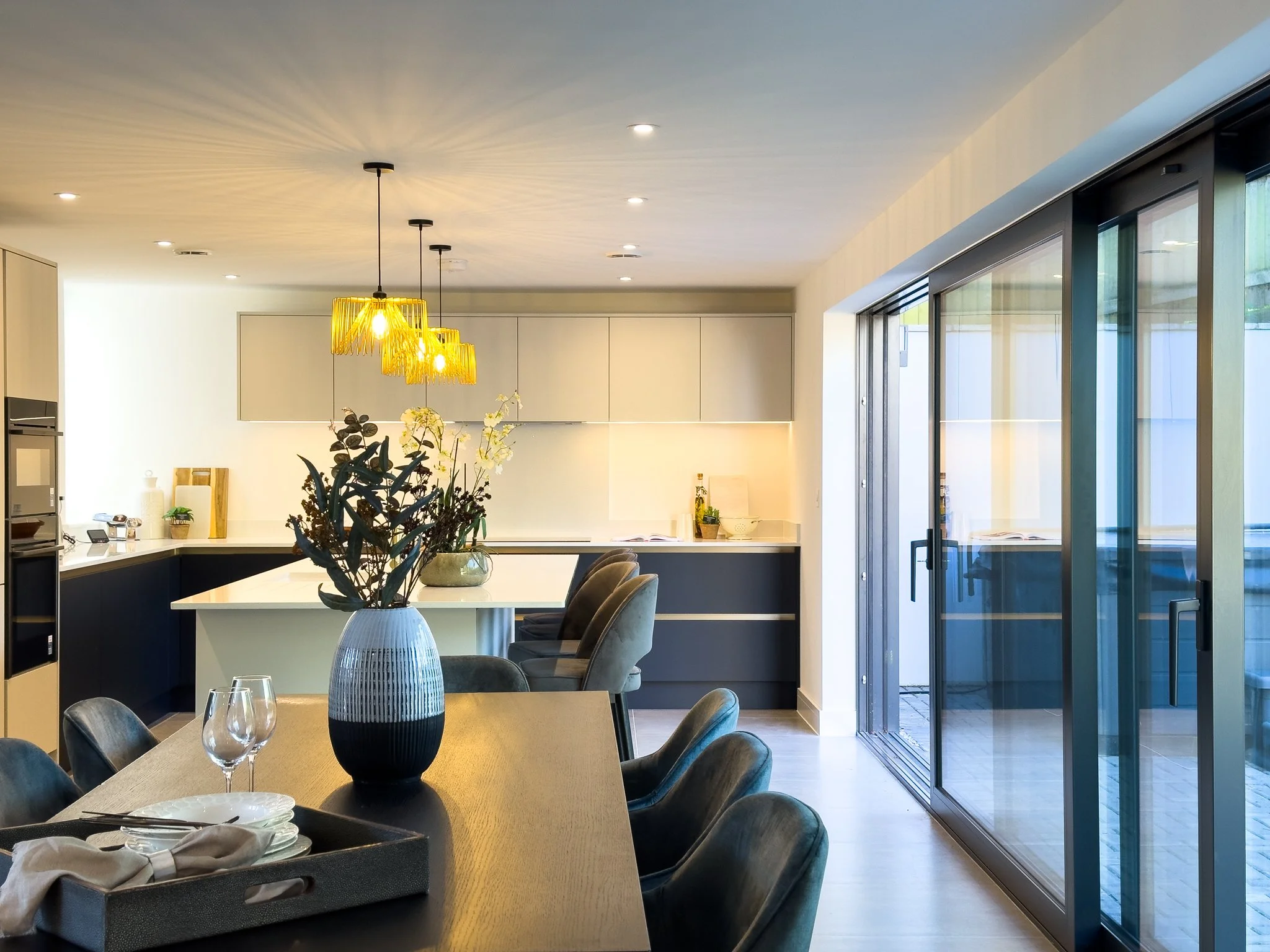 A modern dining room with a large wooden table, black chairs, a blue and black vase with flowers, and a view of an outdoor patio through sliding glass doors. Kitchen in the background with yellow pendant lights.
