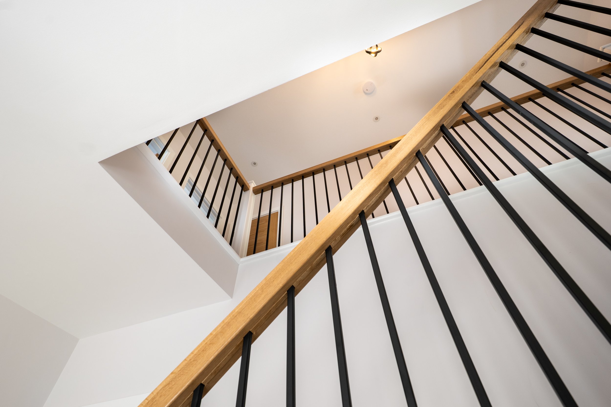View of a staircase from below, featuring wooden handrails and black vertical balusters, with a white ceiling and walls, a ceiling light, and smoke detectors.