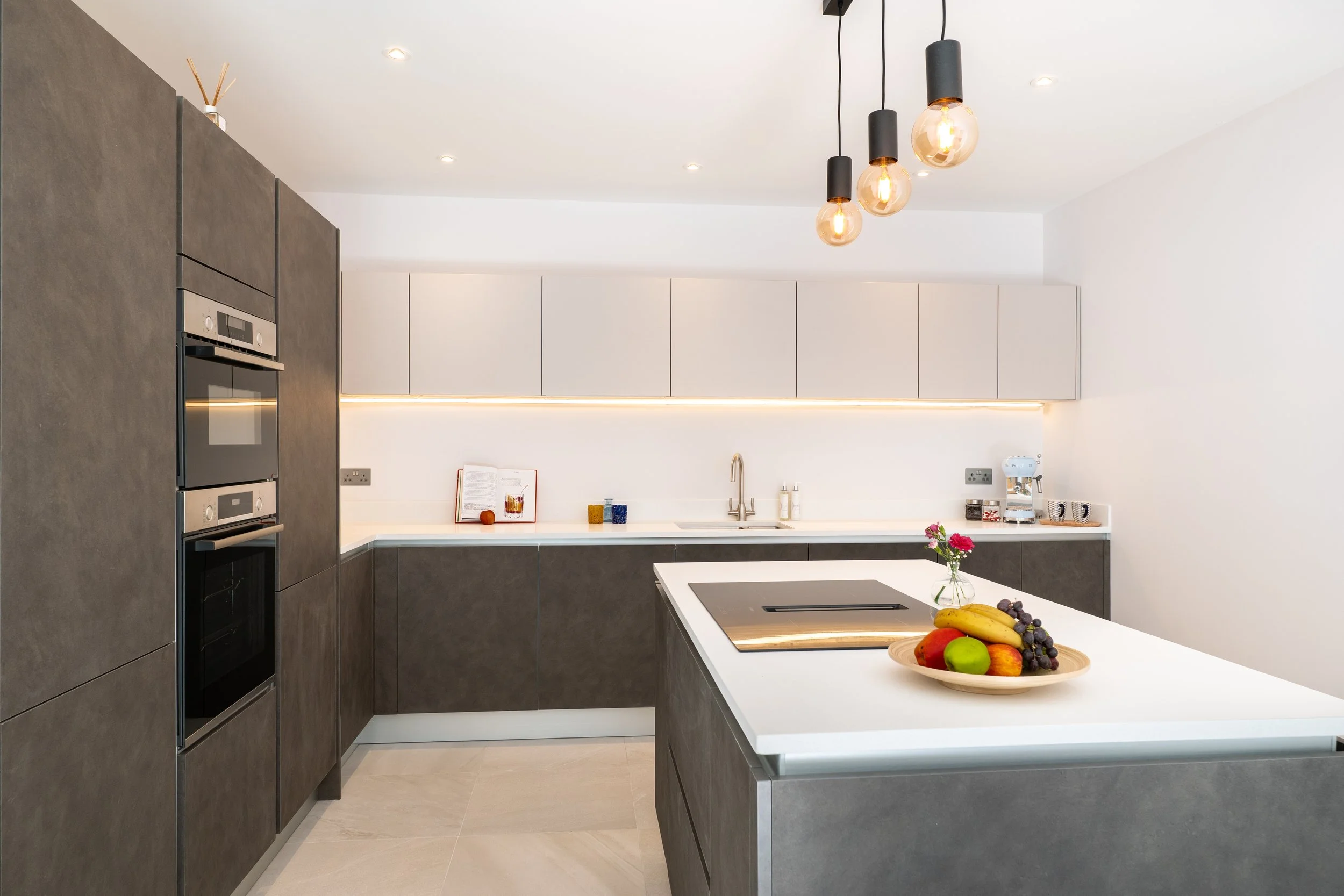 Modern kitchen with white upper cabinets, gray lower cabinets, a white countertop, and pendant lights with exposed bulbs hanging from the ceiling. There is a built-in oven and microwave, a sink with a faucet, and a kitchen island with a bowl of assor