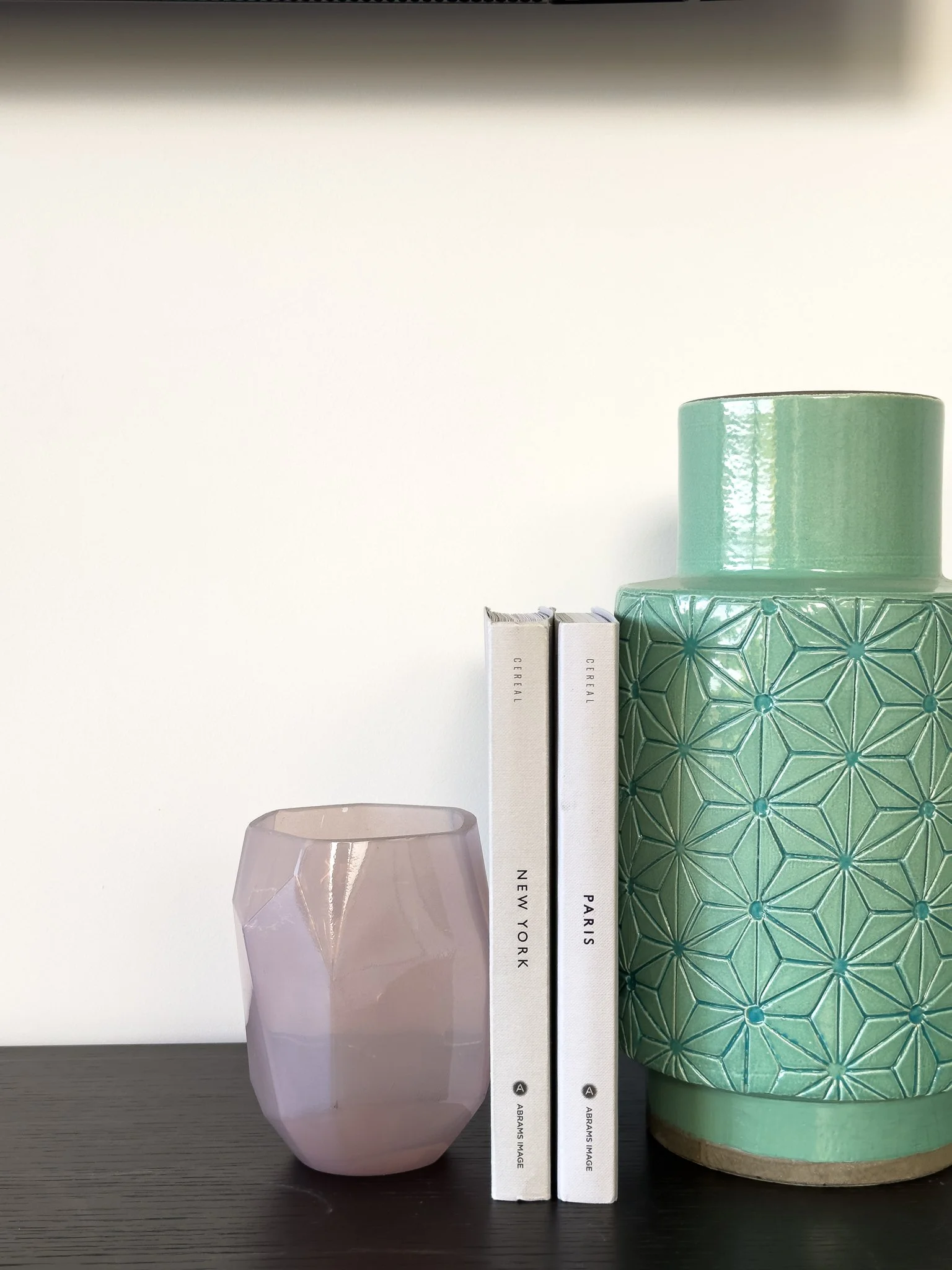 Arrangement of decorative objects on a dark surface including a pinkish closed glass vase, a transparent glass, and two books standing upright with white covers titled 'New York' and 'Paris', with a large green ceramic vase with geometric patterns in