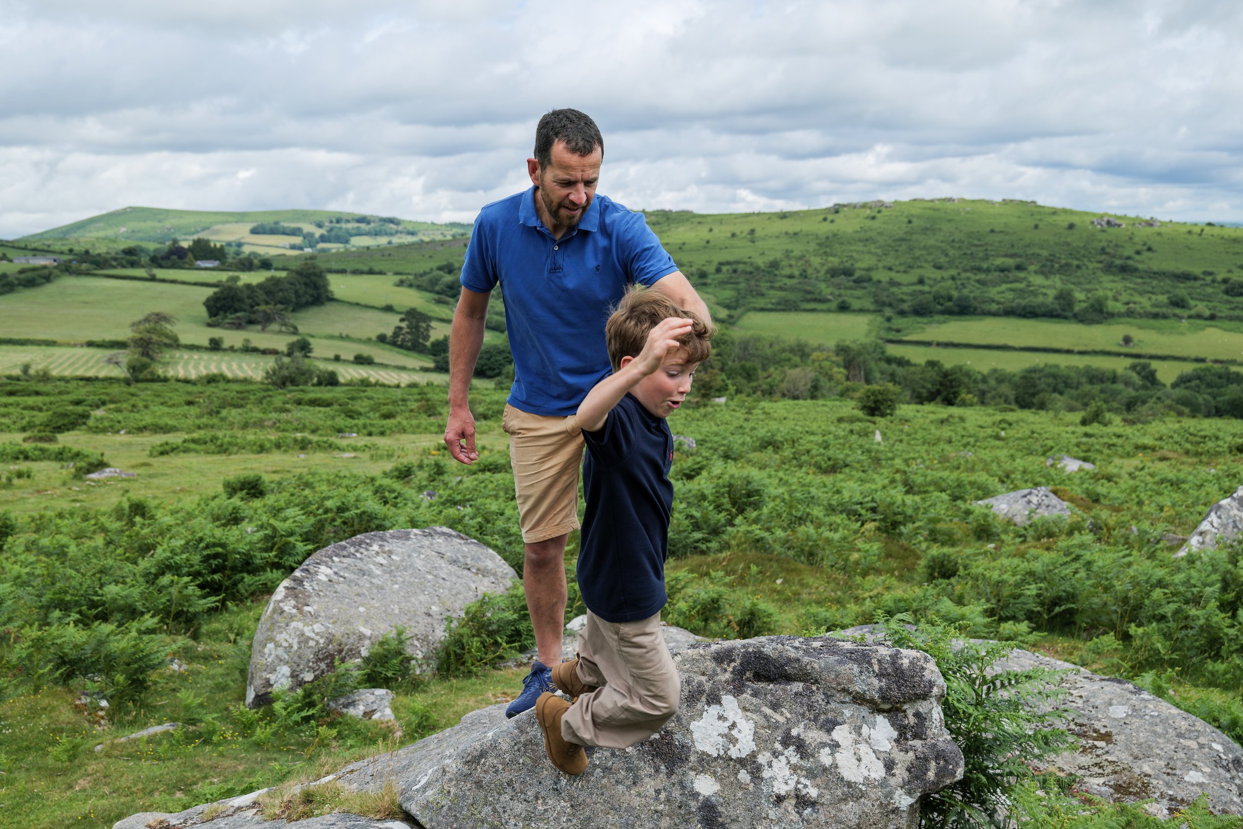 A man and a young boy hiking on rocky terrain in a green countryside with rolling hills and cloudy sky.