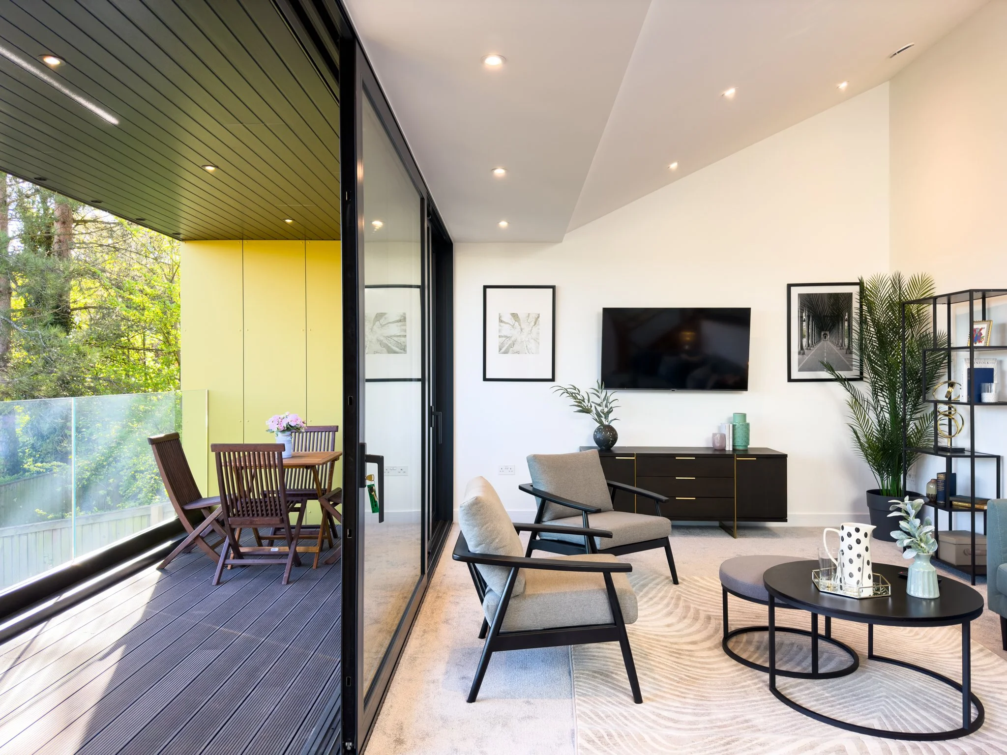 Modern living room with a large flat-screen TV, framed artwork, and a round coffee table decorated with vases and glasses. A comfortable armchair, a shelf with books and plants, and a sliding glass door leading to a balcony with outdoor seating and trees outside.