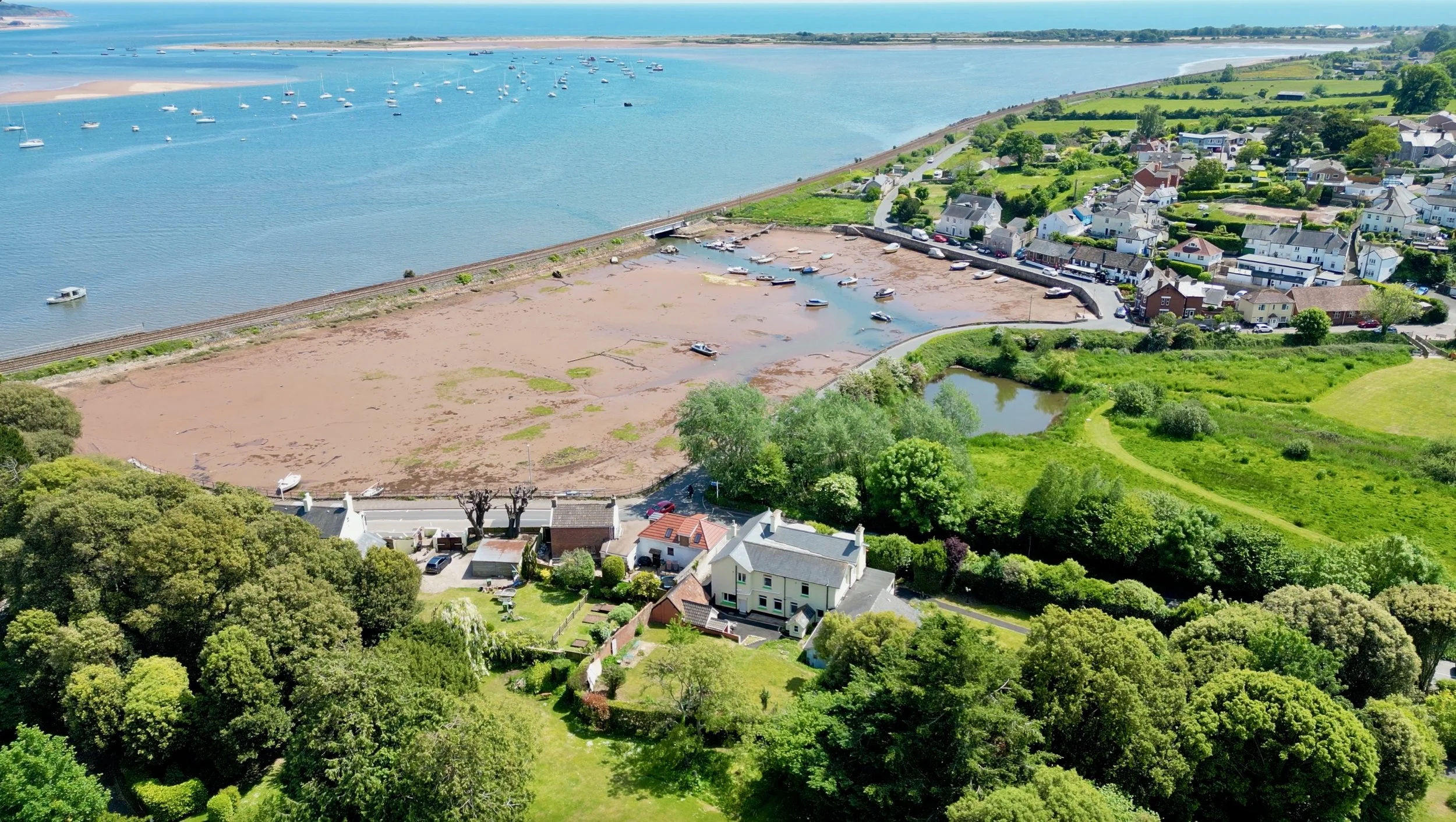 A coastal residential area with houses surrounded by lush green trees, adjacent to a beach and a body of water with boats anchored, and a sandbar or tidal area with boats grounded during low tide.