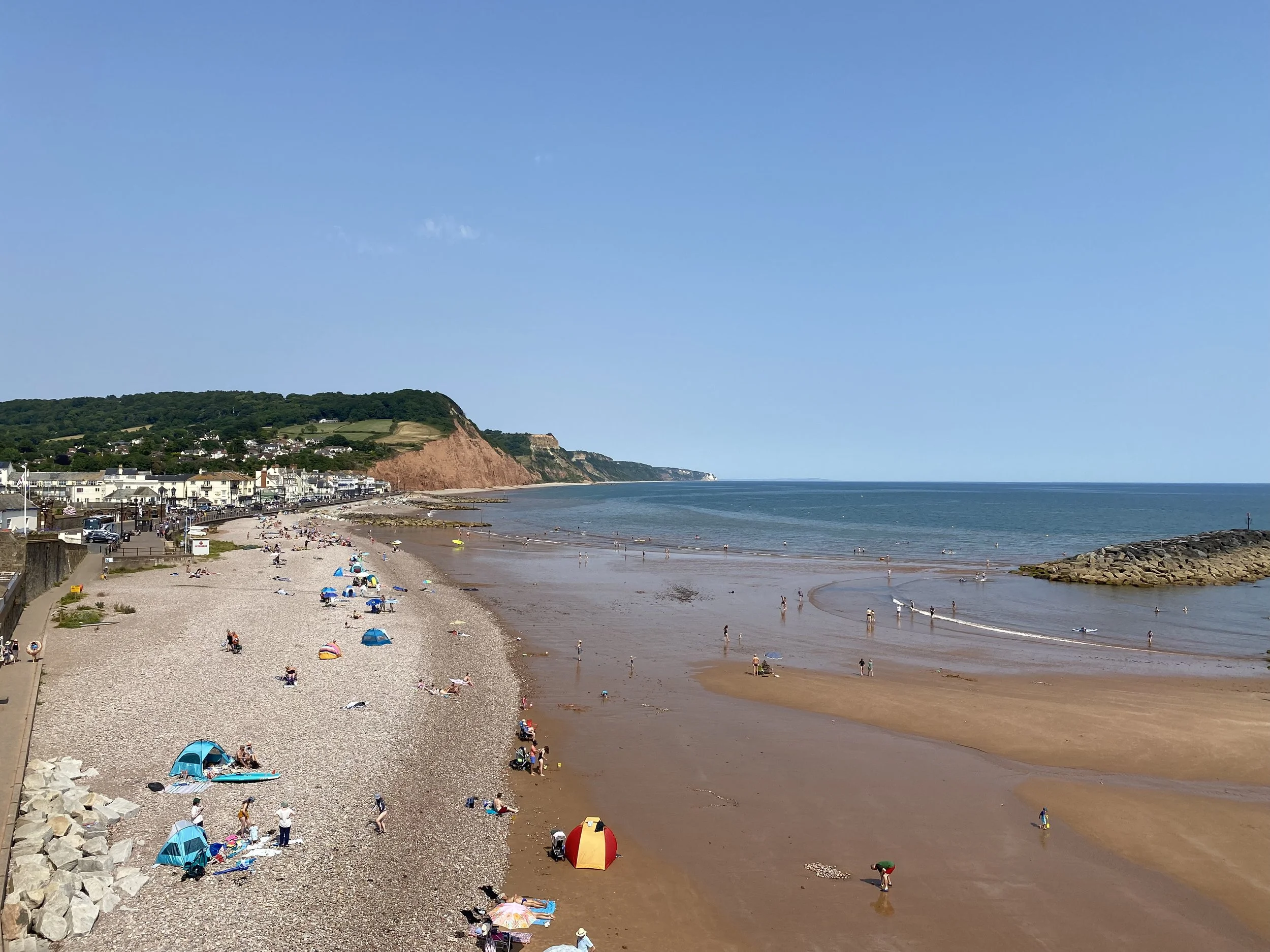 A beach scene with people relaxing and playing near the shoreline, colorful umbrellas and tents on the sandy beach, a rocky shoreline, a green hillside with houses, and cliffs in the background under a clear blue sky.