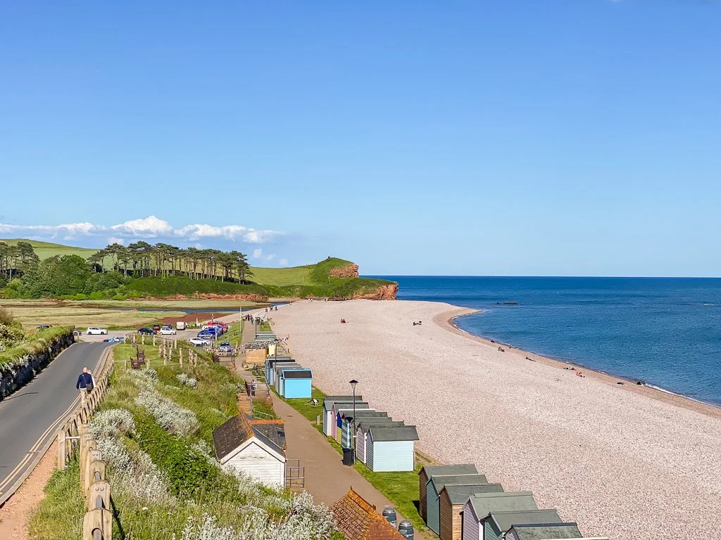 A coastal beach with sand and a few people, green hills with trees in the background, a road with cars and a person walking, and colorful beach huts along the shoreline.