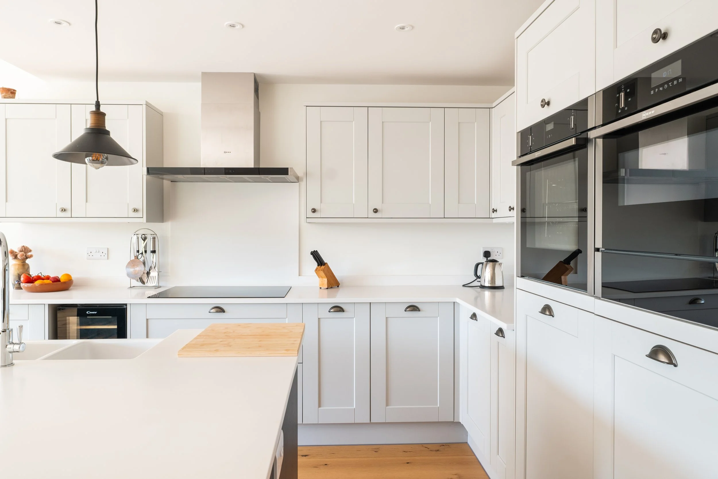 Modern white kitchen with built-in appliances, a black pendant light, a bowl of fruit, and various kitchen tools on white countertops.