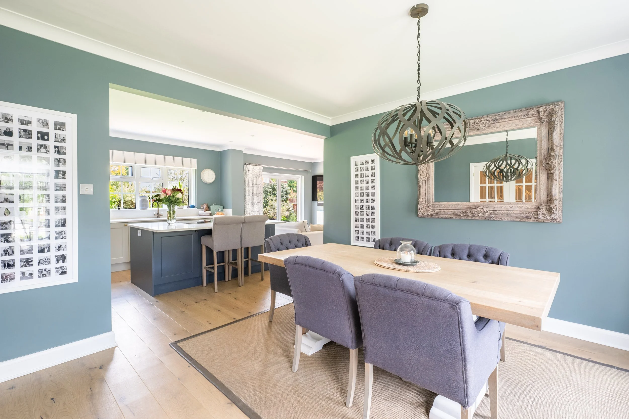 Open-concept living and dining area with blue walls, a wooden dining table, upholstered chairs, a large ornate mirror, pendant lighting, and a view into the kitchen with a vase of flowers on the counter.