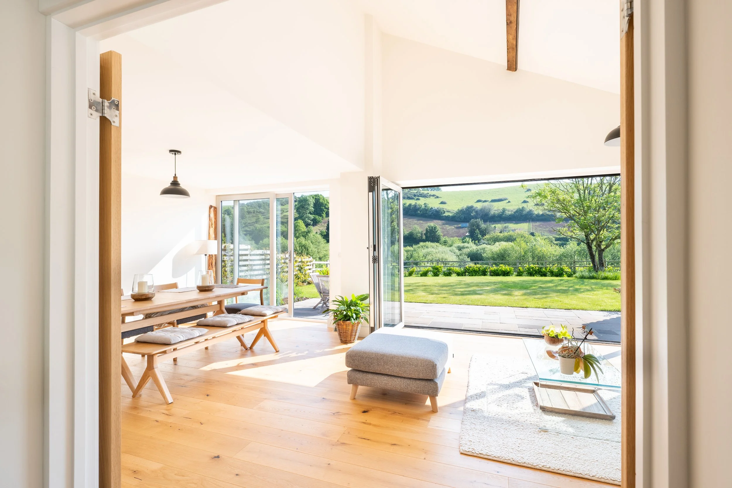 Bright, airy living space with an open door leading to a lush, green outdoor landscape with trees, grass, and rolling hills. Inside, there is a minimalist wooden dining table with cushions, a gray ottoman, and potted plants, with natural light floodi