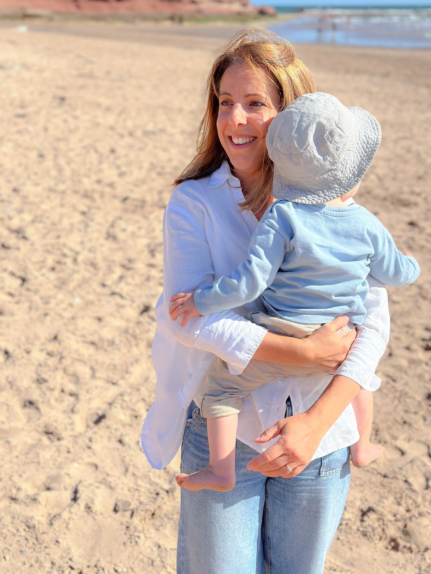 A woman holding a young child on a sandy beach with waves and a clear sky in the background.