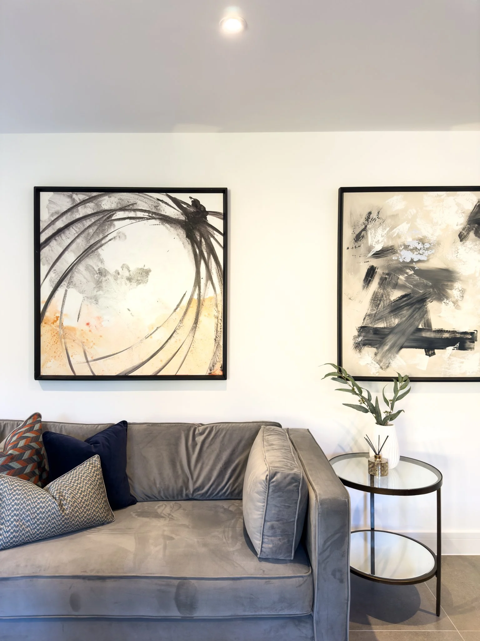 Modern living room with a gray sofa, various patterned pillows, two black and white abstract paintings on the wall, a potted plant in a white vase, and a black side table with a glass top.