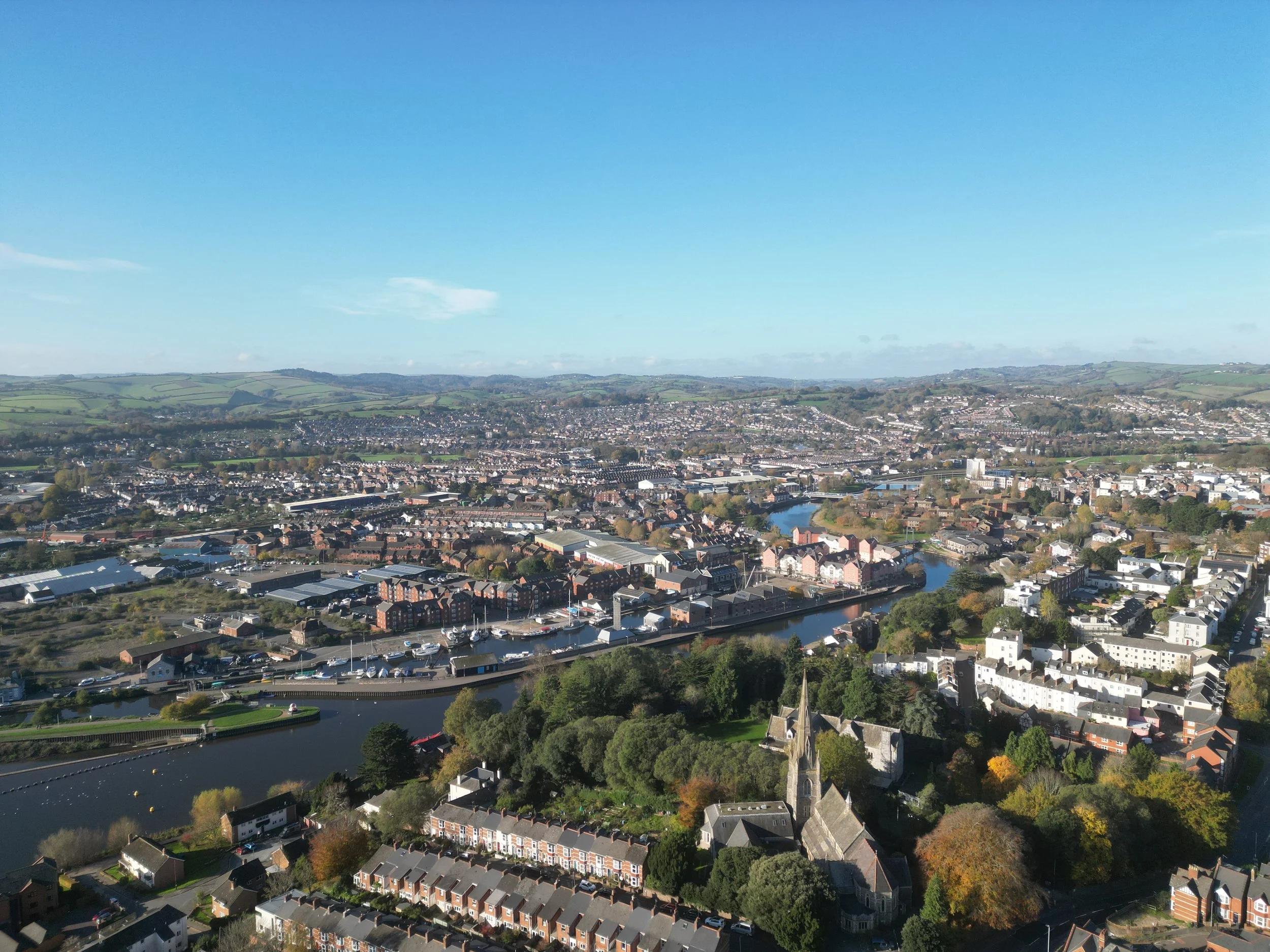 Aerial view of a city with a river, residential buildings, and green hills in the background under a blue sky.