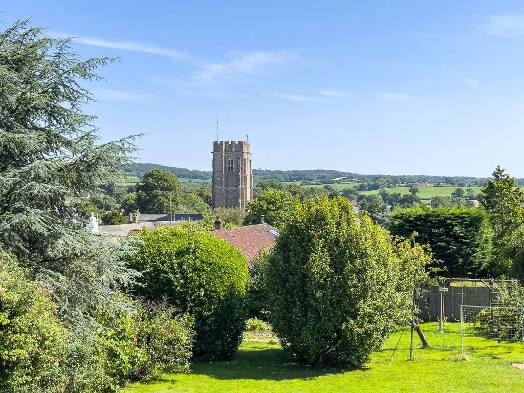 A scenic view of a small town with a prominent stone church tower in the background, surrounded by lush green trees and grass, under a clear blue sky.