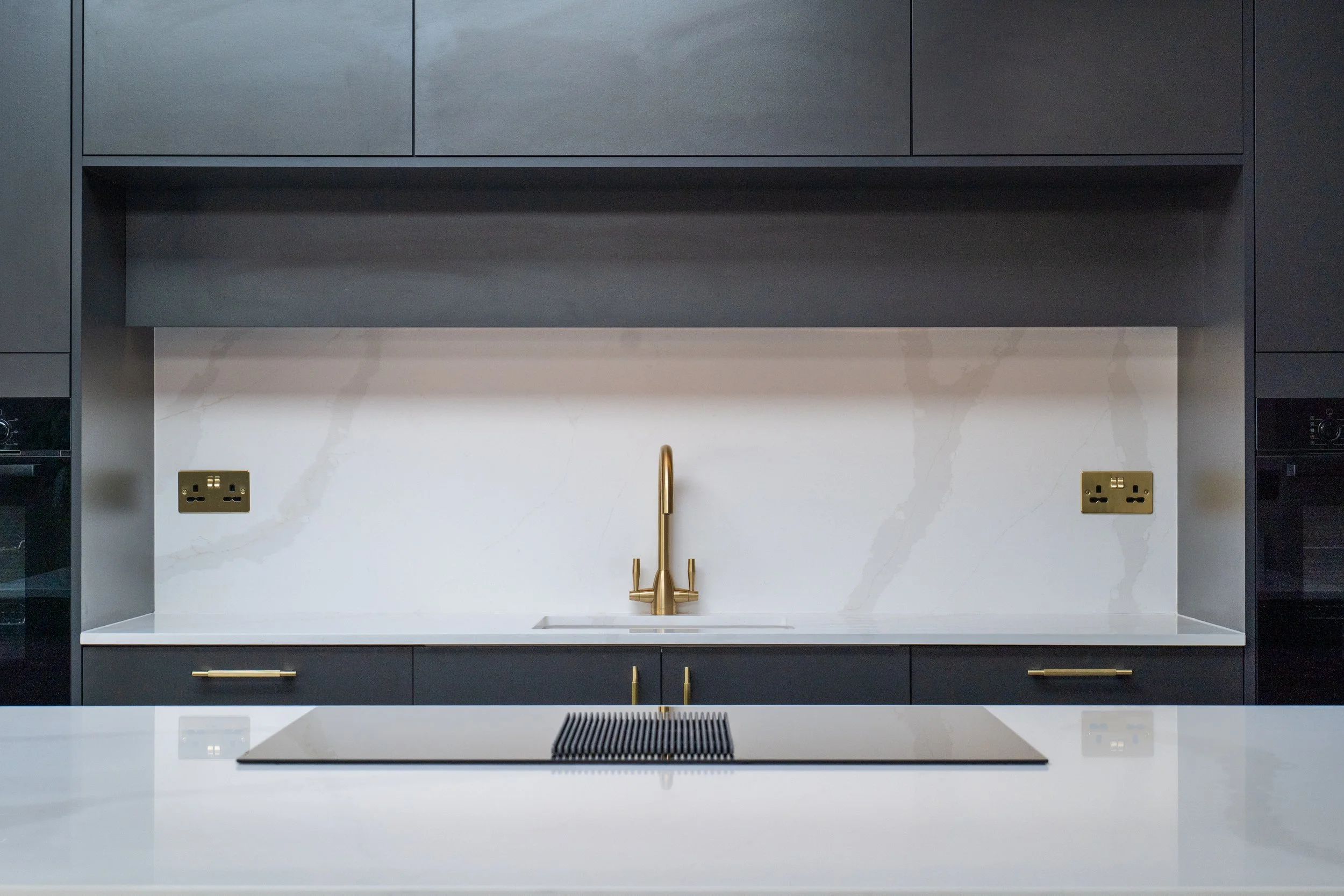 Modern kitchen with black cabinets, white marble backsplash, gold faucet, and two outlets.
