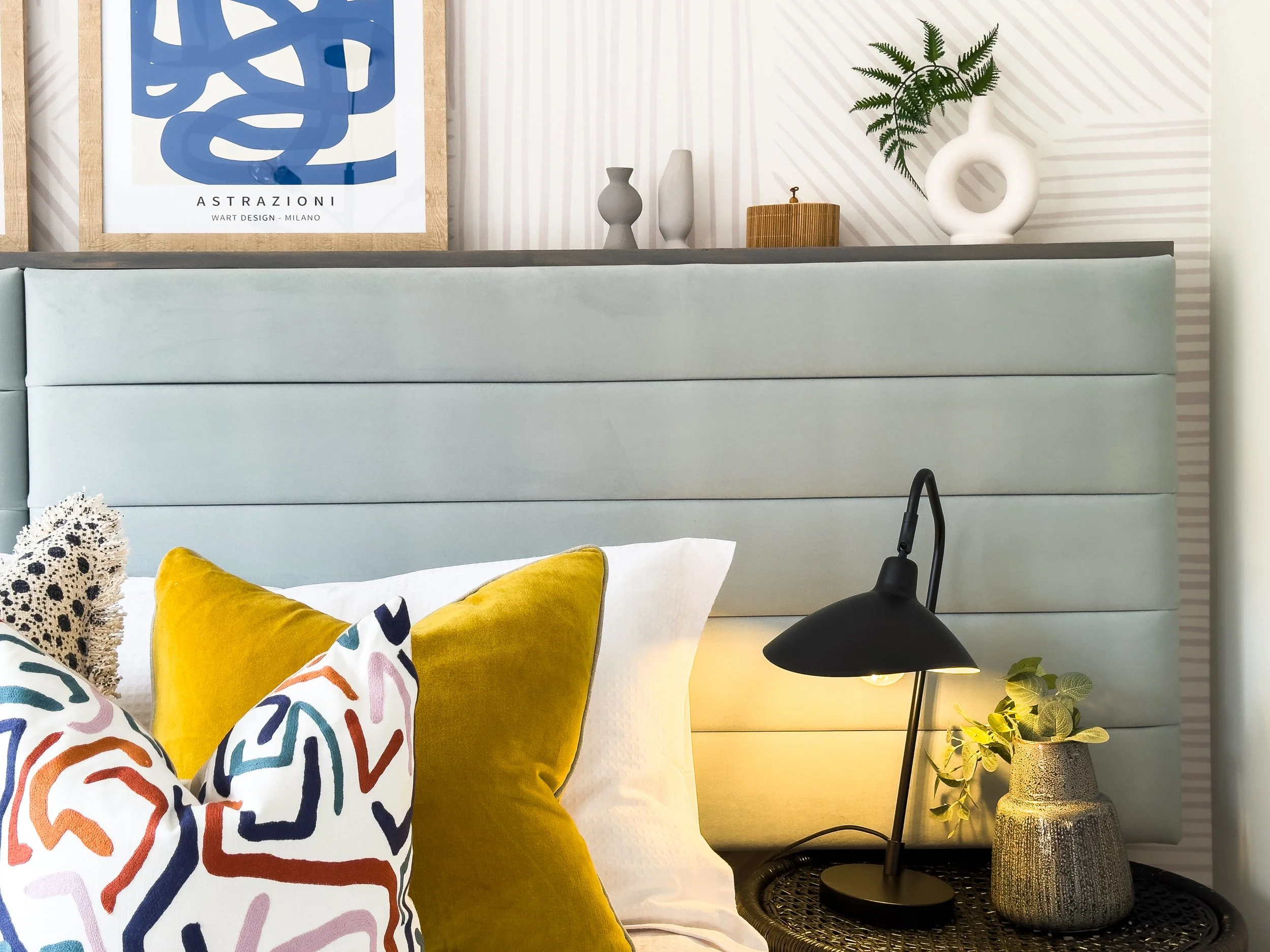 A bedroom decor scene featuring a light blue upholstered headboard, decorative pillows in yellow and multicolored patterns, a black table lamp, and a tall textured vase with green foliage on a side table. There is a framed blue abstract art piece wit