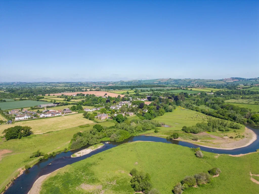 Aerial view of a lush green landscape with a winding river, fields, trees, and a small cluster of houses under a clear blue sky.