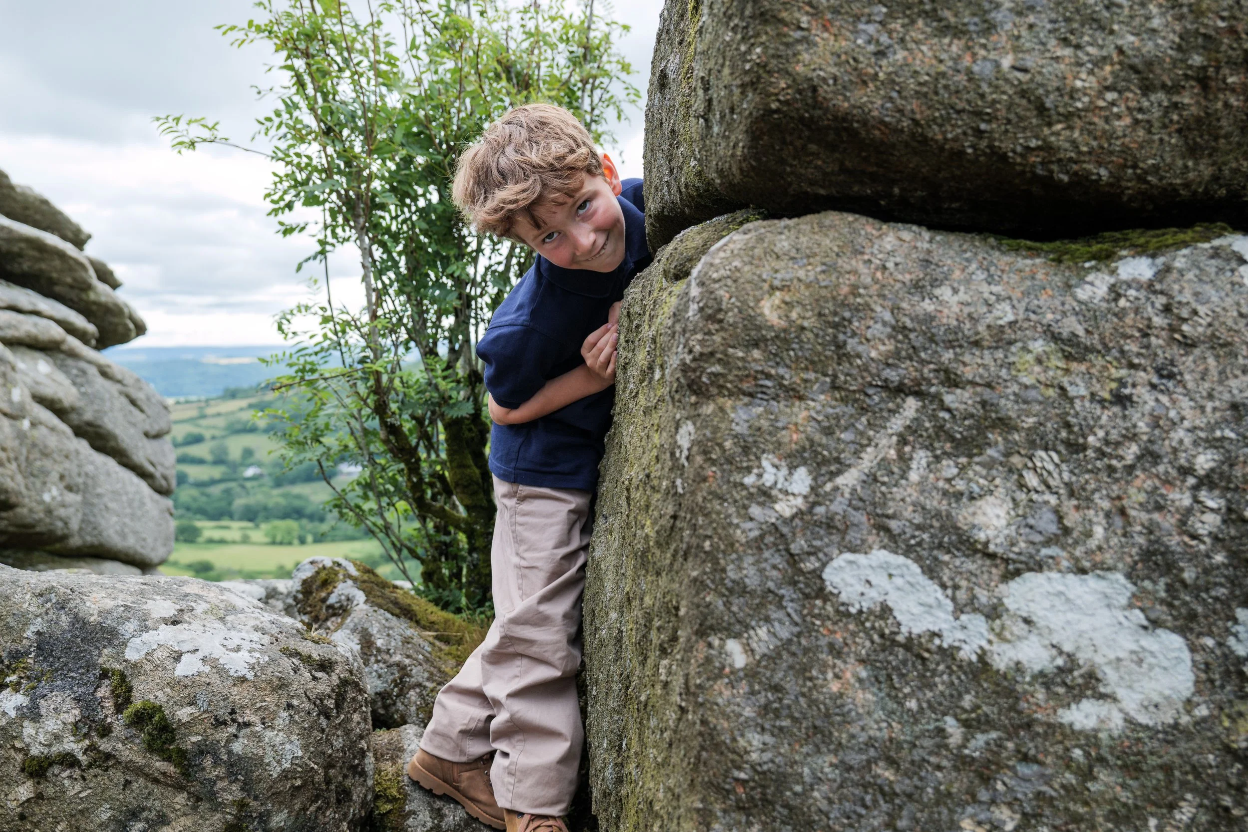 A young boy with curly hair peeking around a large stone wall outdoors, with a scenic green landscape and cloudy sky in the background.
