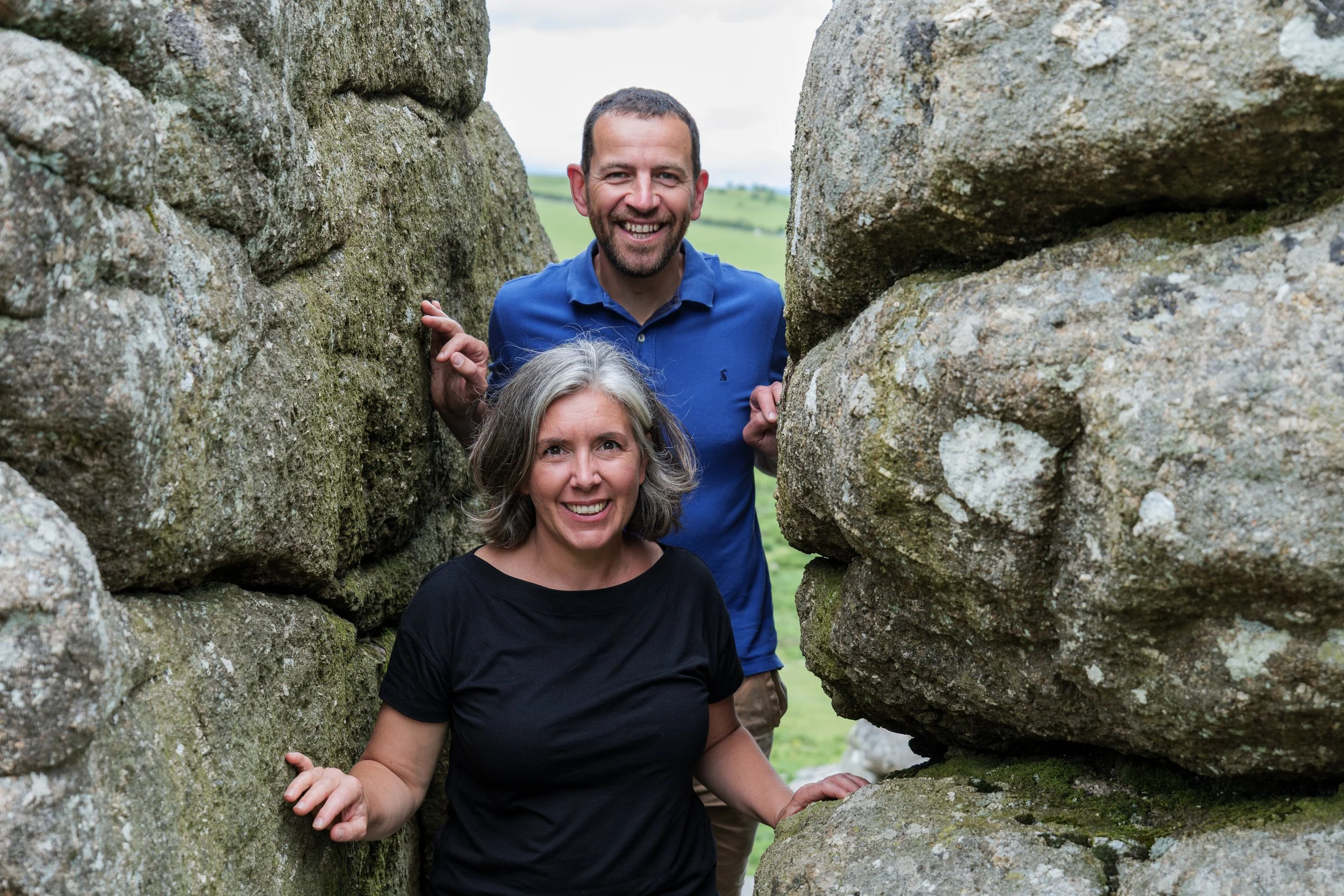 Two smiling people, a man and a woman, peeking through a gap between large rocks, with a green field and cloudy sky in the background.