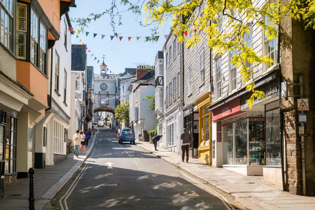 A street scene in a small town with shops and pedestrians, featuring colorful bunting hanging across the street, trees, and buildings with storefronts.