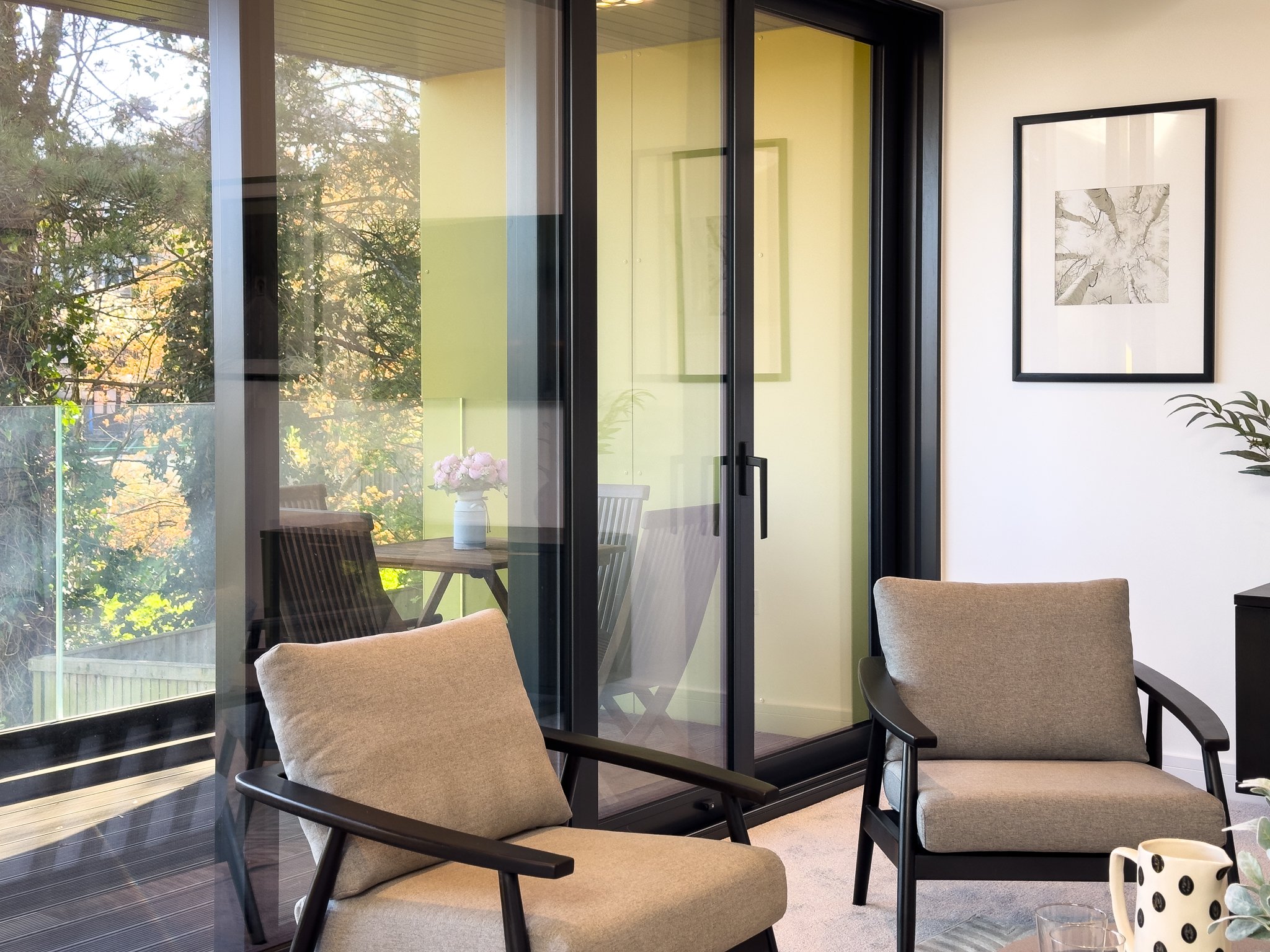 Interior of a modern room with beige chairs, a coffee mug with black dots, and a glass sliding door leading to an outdoor balcony with trees.