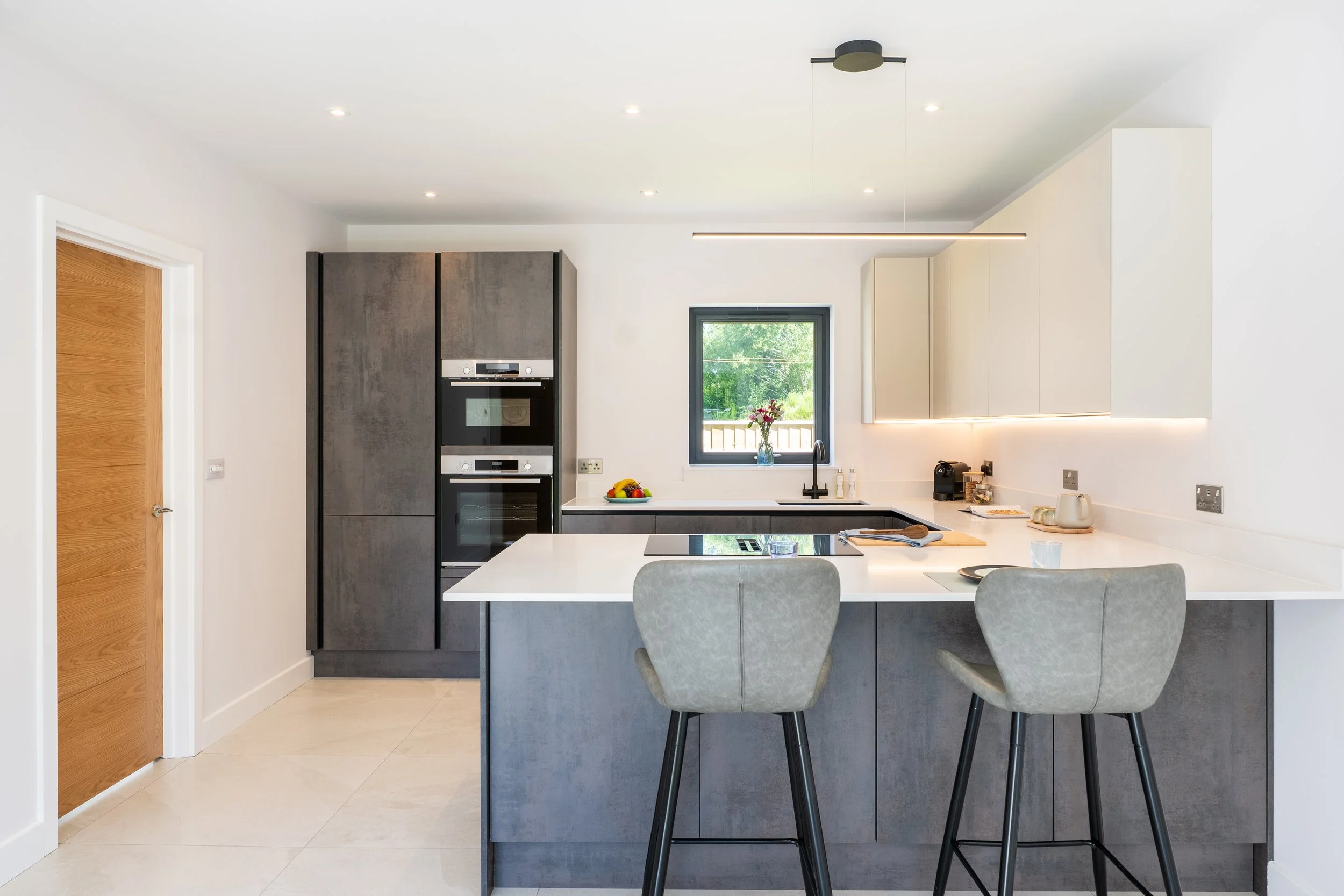 Modern kitchen with white countertops, gray cabinets, black appliances, a window with a view of greenery, and bar stools around the island.