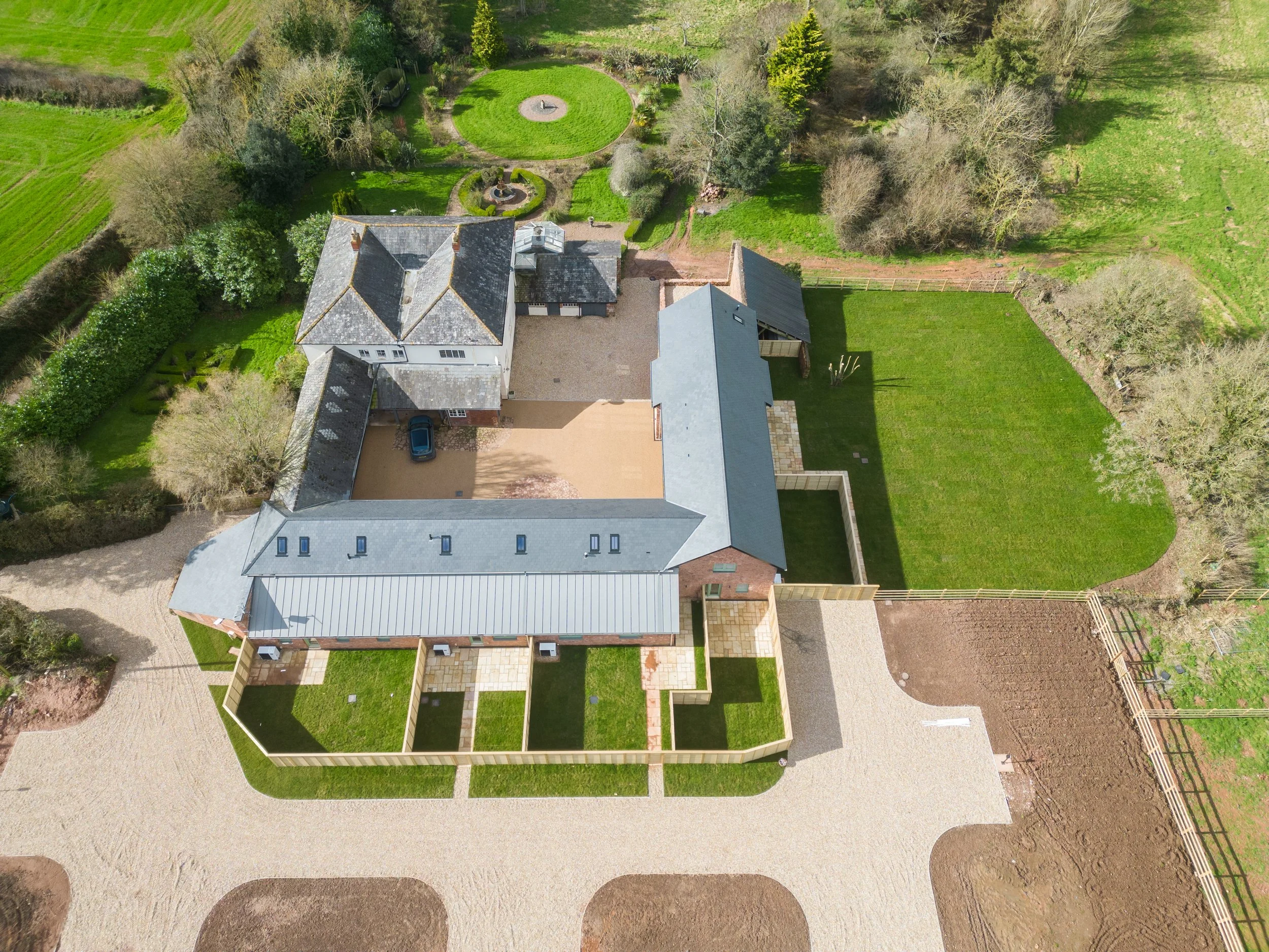 An aerial view of a large house with a gray roof, surrounded by a manicured lawn, small fenced garden, and gravel pathways, with trees and green fields in the background.