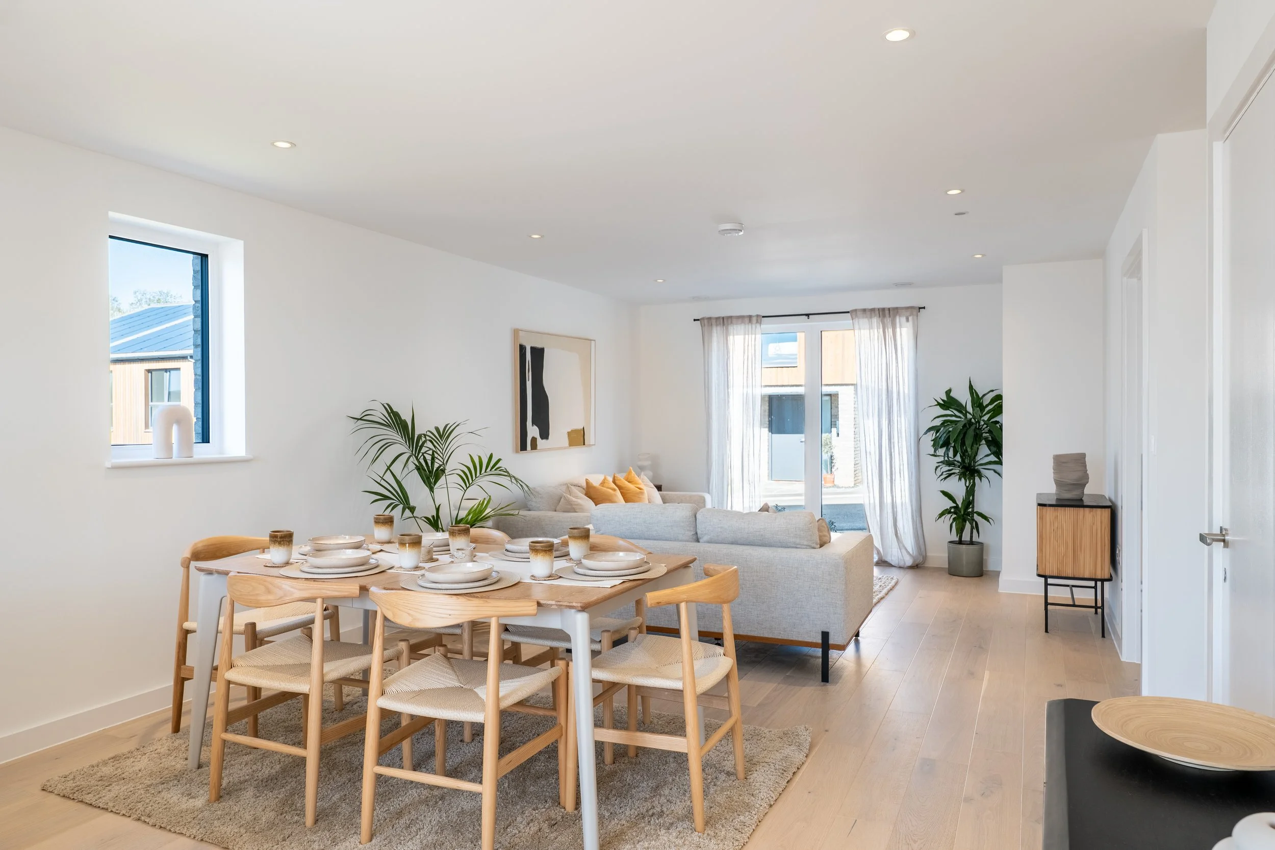 Open living and dining area with large windows, white walls, wooden flooring, a dining table set with plates and glasses, a beige sofa, potted plants, artwork, and curtains.