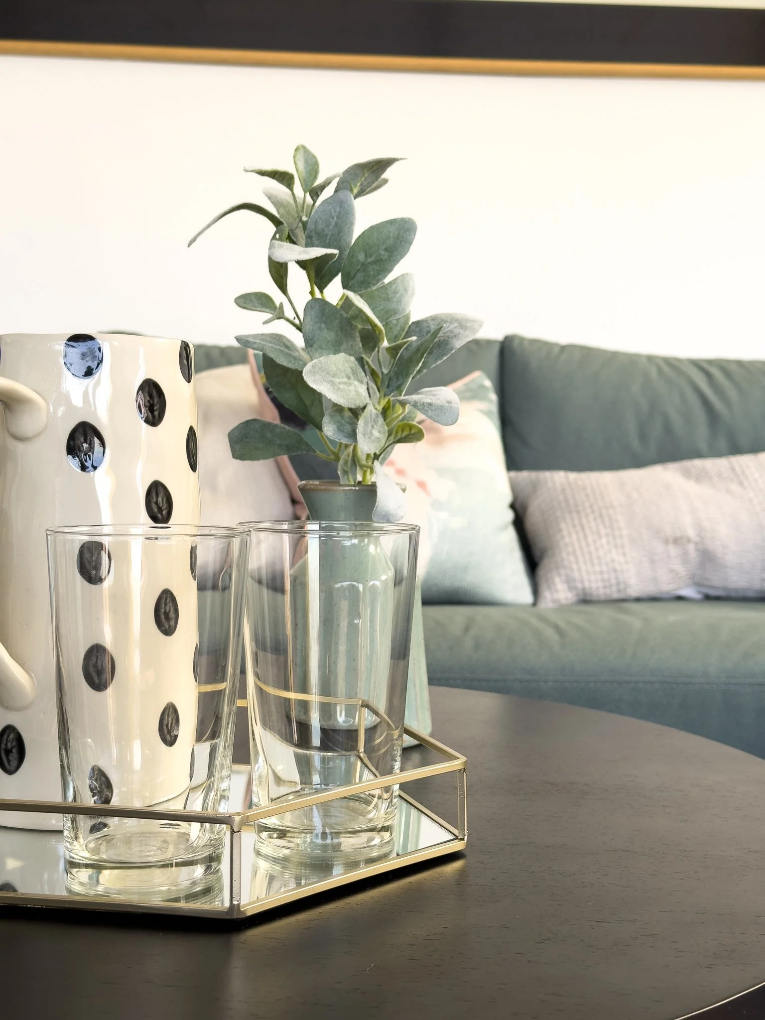 A small potted plant with silvery-green leaves on a table, with empty glasses in the foreground, a polka dot mug, and a contemporary sofa with pillows in the background.
