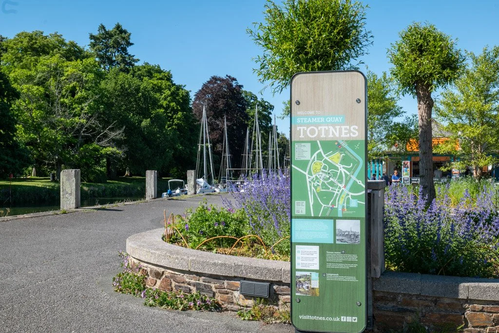 A signboard welcoming visitors to Steamer Quay in Totnes, with a map and information. There are trees and purple flowers in the foreground, and boats docked at the quay in the background.