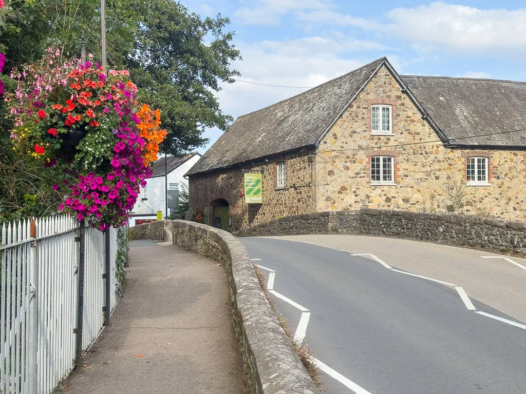 A street scene featuring a curved road with a stone wall on one side and a sidewalk with a colorful flower arrangement hanging from a fence. There is a stone building with small windows and a sloped roof in the background, under a partly cloudy sky.