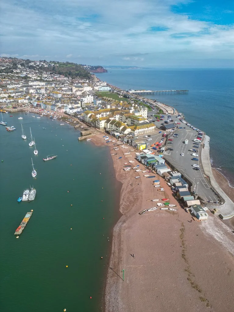 A coastal town with a harbor and boats, a sandy beach with small boats, a parking lot, and residential buildings along the shoreline.