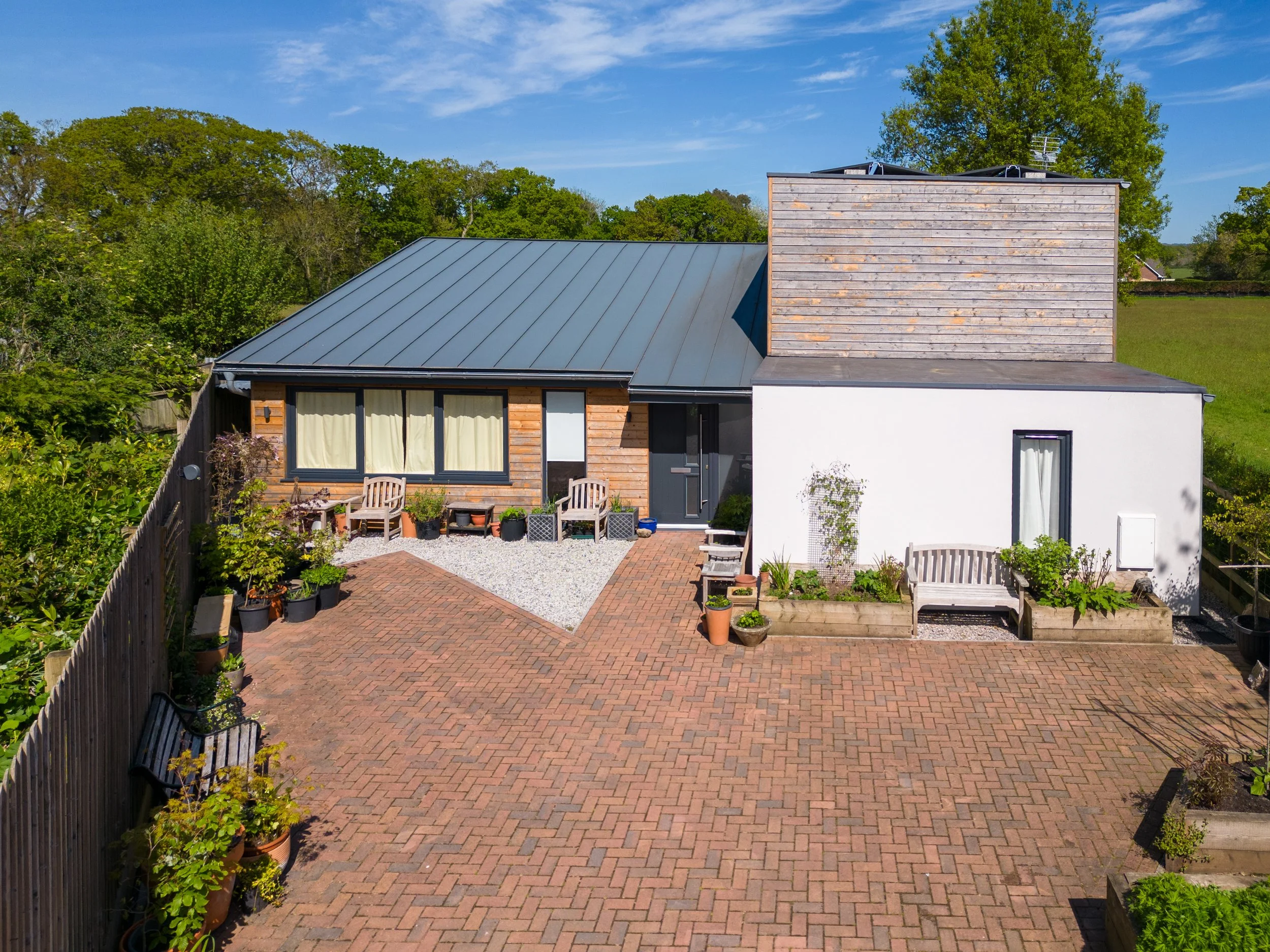 Rear view of a modern house with brick patio, potted plants, and garden furniture, surrounded by trees and greenery under a blue sky.