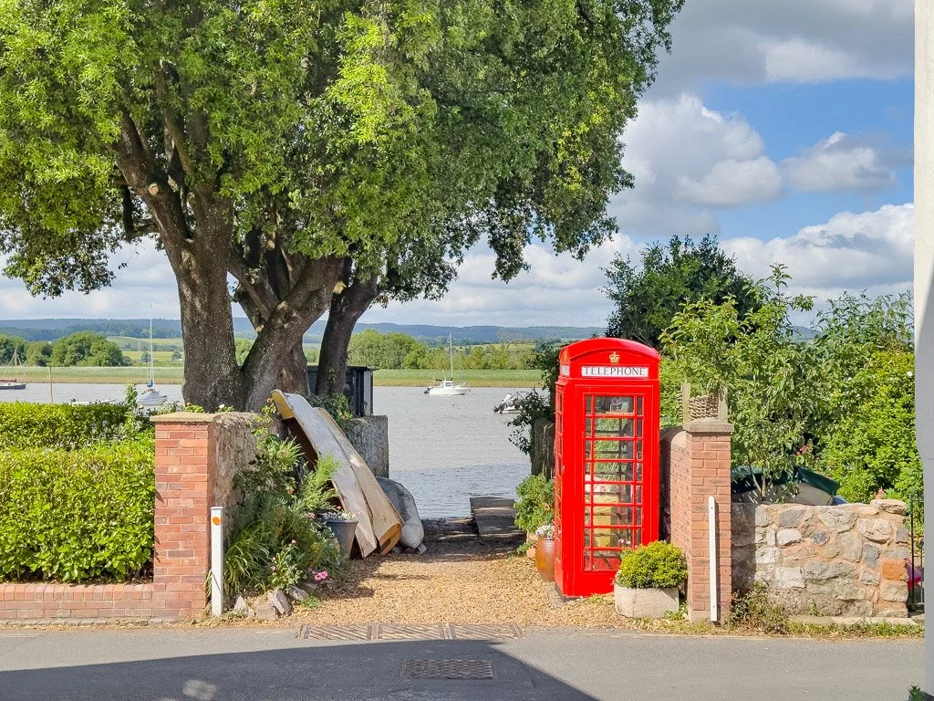 A red British telephone booth located on a gravel path near a body of water with boats, surrounded by trees and bushes, with a brick and stone wall, and a large tree providing shade.