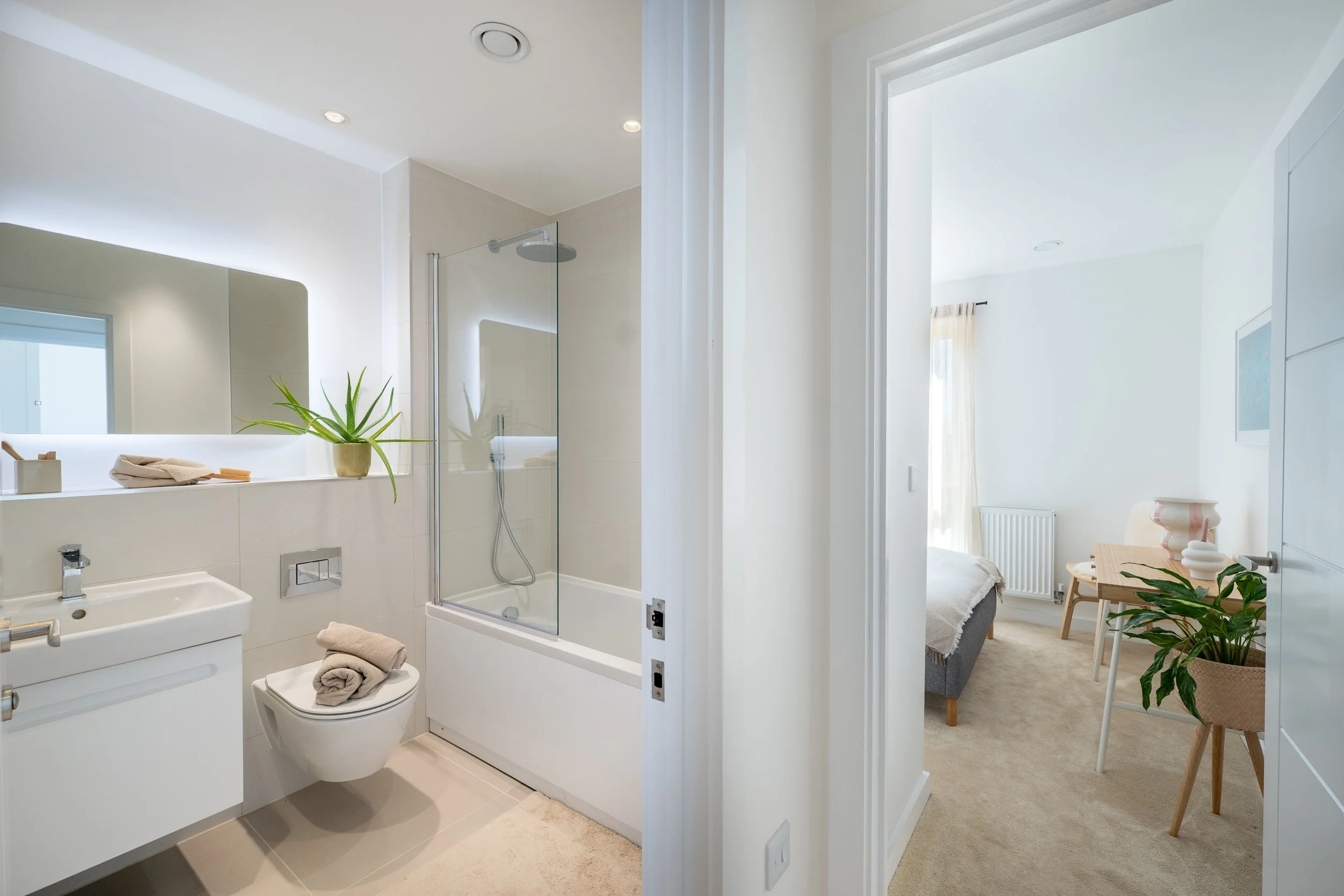 A modern bathroom with a shower, toilet, and sink, decorated with a potted plant and towels. An open door reveals a glimpse of a bedroom with a bed, a chair, and a window with curtains.