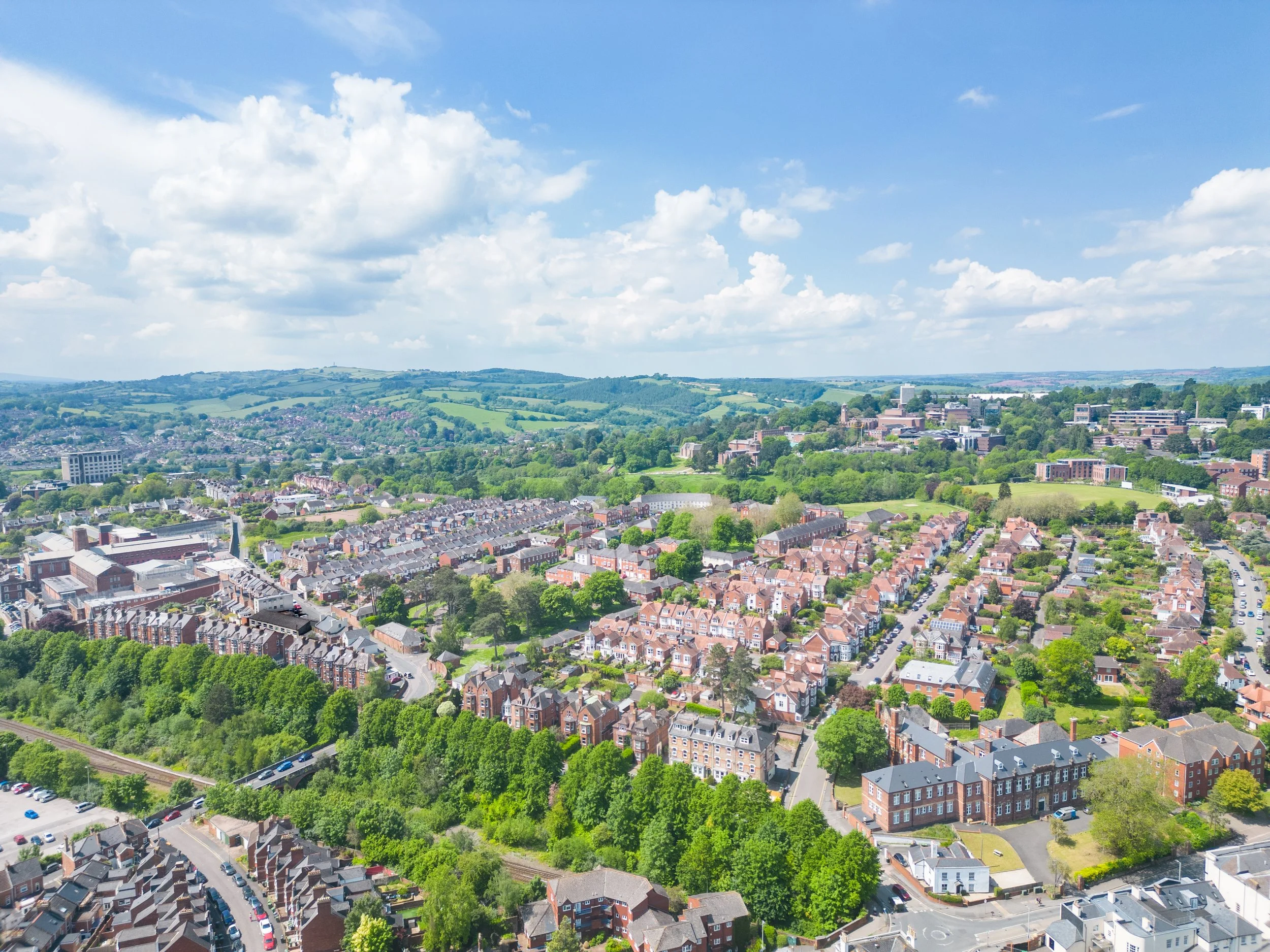 Aerial view of a city surrounded by green hills and trees, with residential houses and a few commercial buildings under a partly cloudy blue sky.