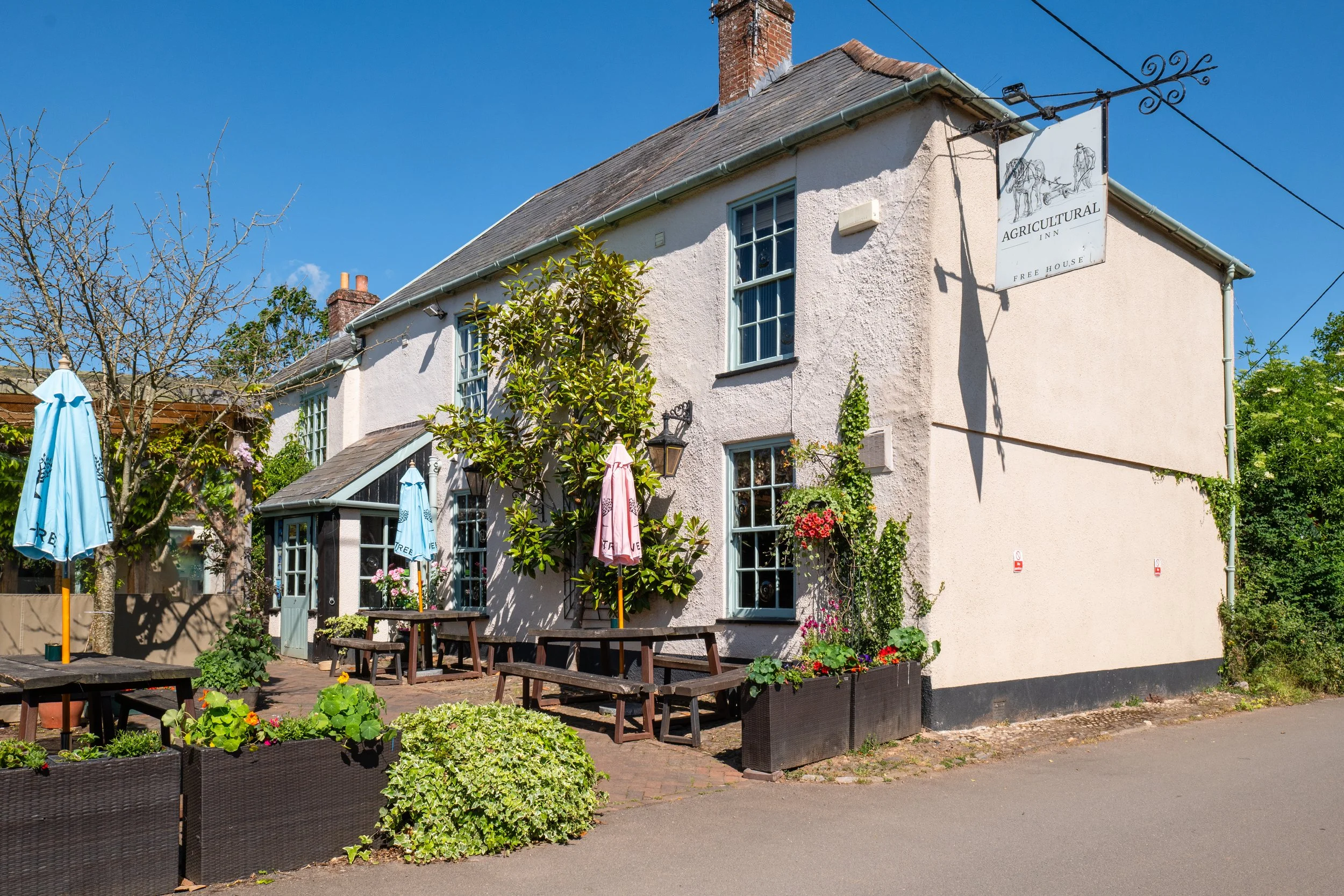 Exterior of a white building with a sign that reads 'Agricultural Inn Free House' and features a drawing of a person and horse-drawn carriage. Outdoor seating with umbrellas, wooden benches, and planters with flowers and greenery.
