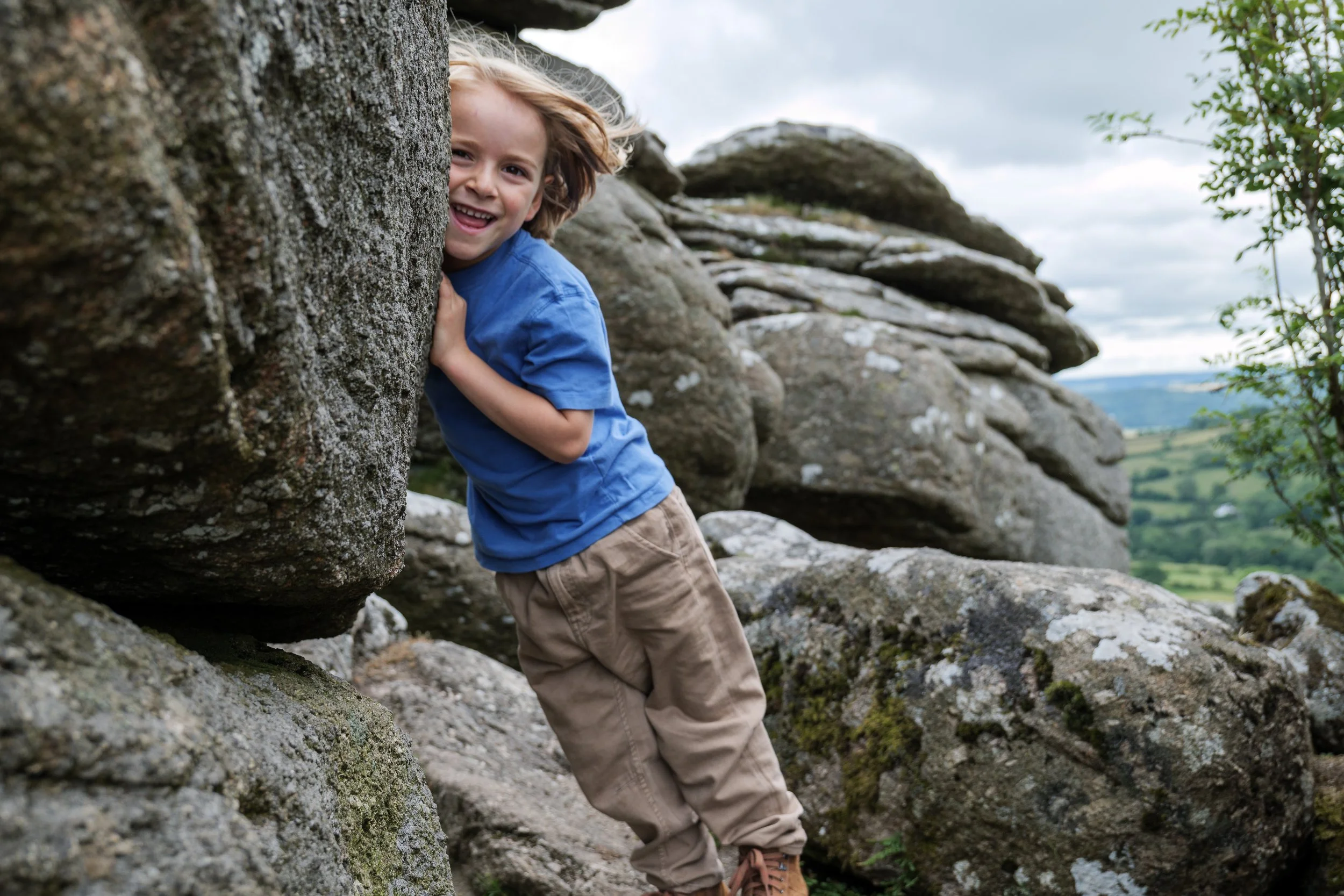 A young boy with long hair wearing a blue shirt and beige pants is smiling while peeking out from behind a rocky outdoor terrain with a cloudy sky in the background.