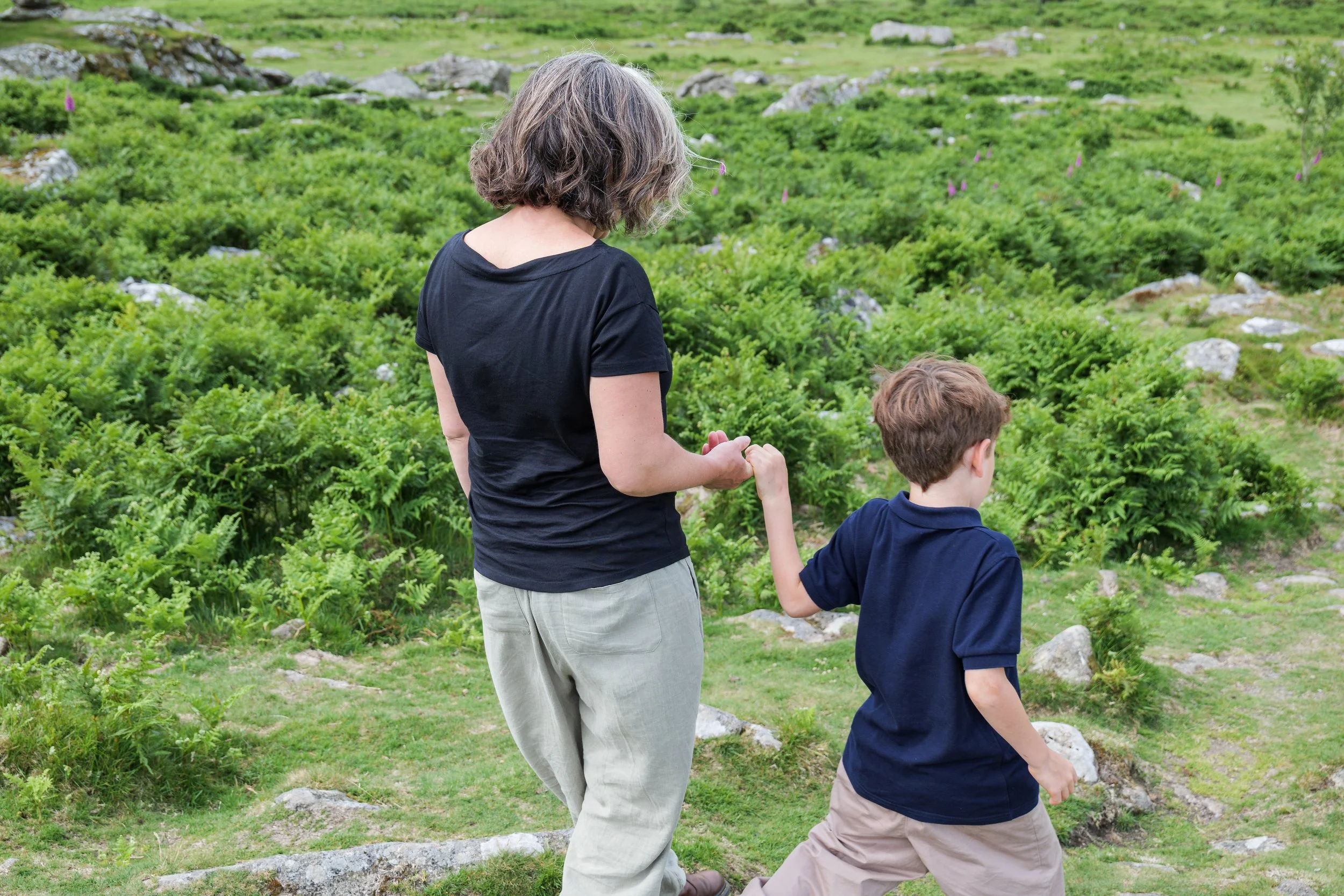 A woman with short gray hair in a black t-shirt holding hands and walking with a young boy in a navy blue shirt and khaki pants through a green, rocky meadow with plants and rocks in the background.