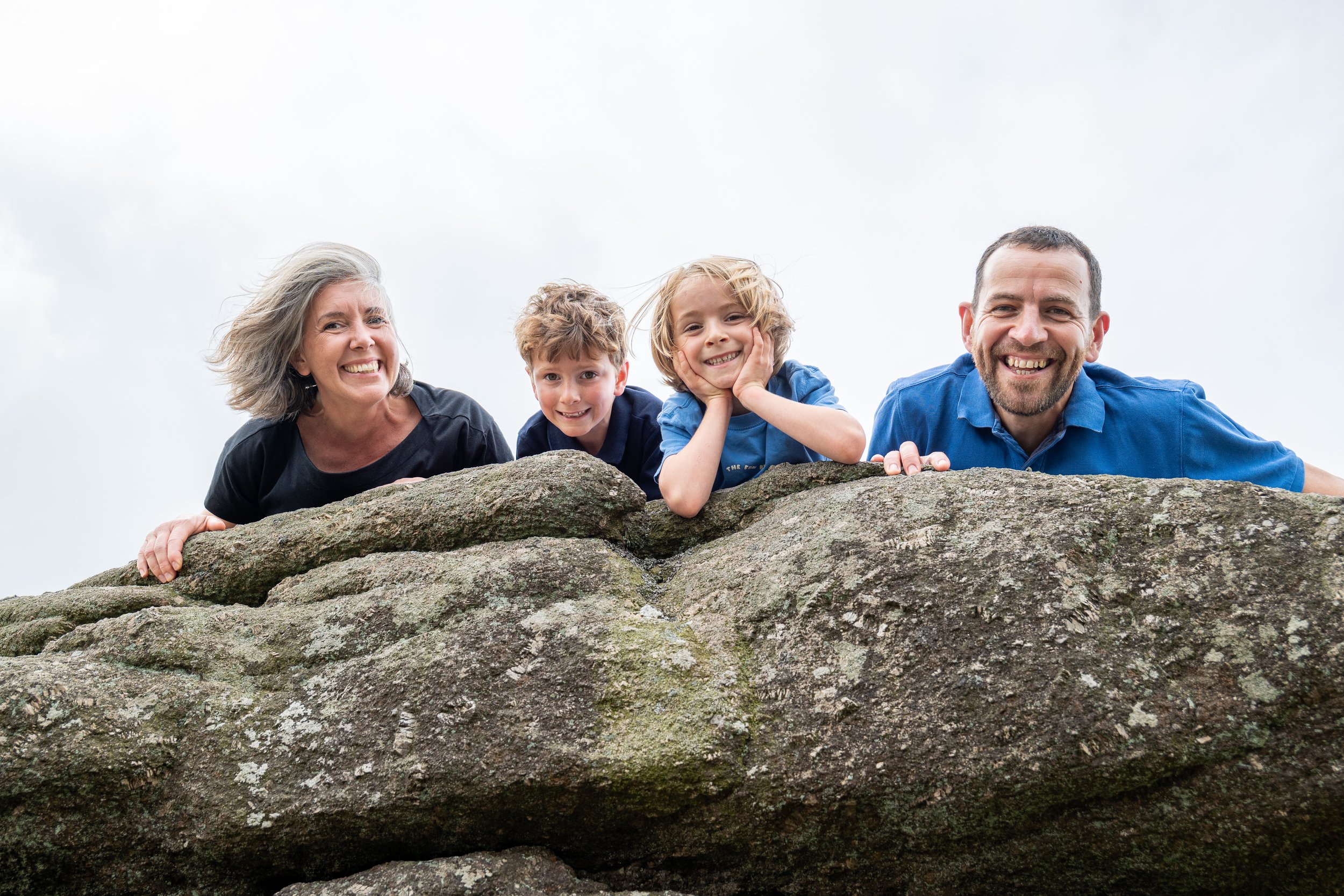 A family of four lying on rocks outdoors, smiling at the camera with cloudy sky background.