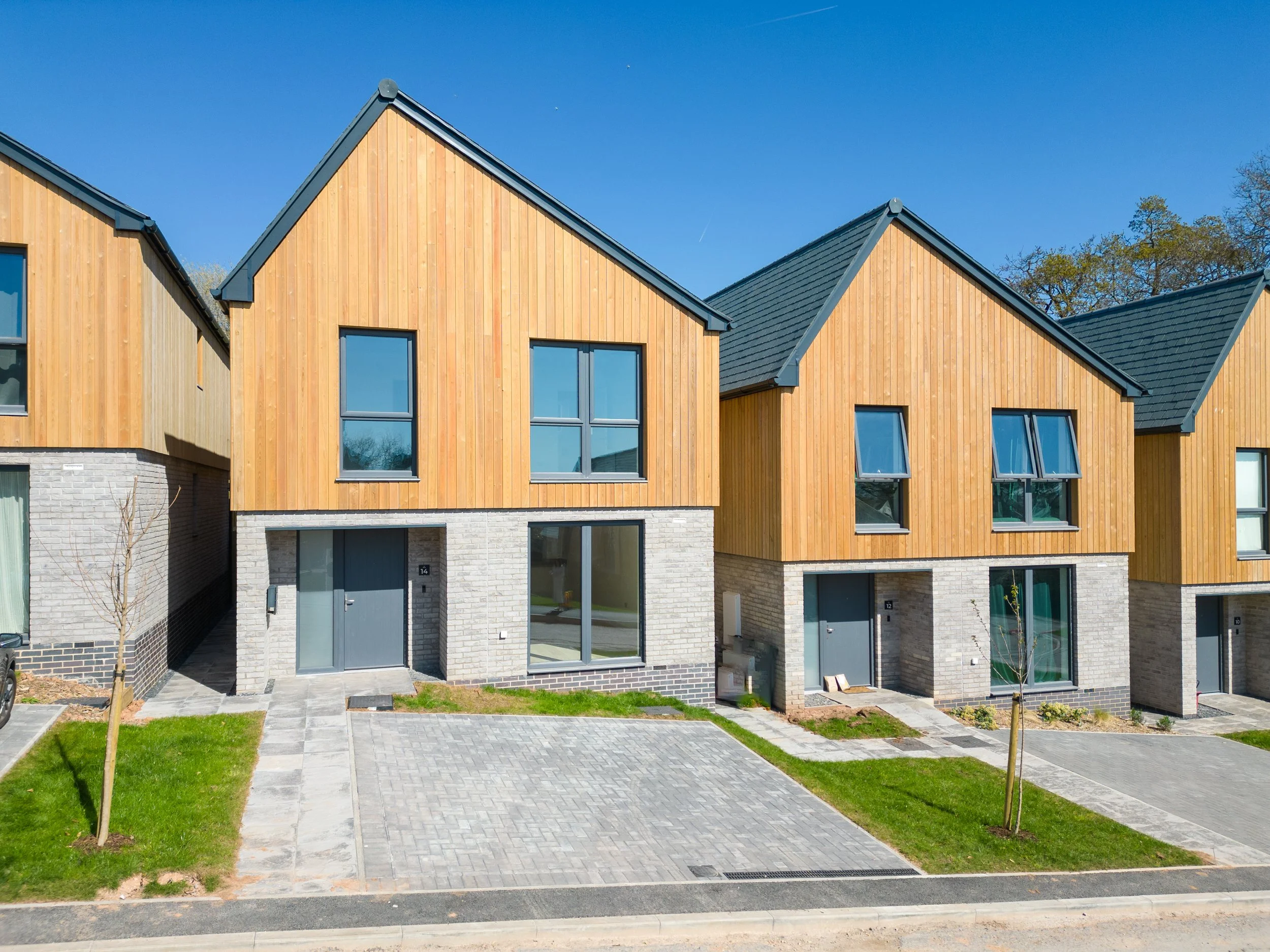 Modern townhouse with wooden siding, large windows, and a paved front yard with grass and small trees under a clear blue sky.