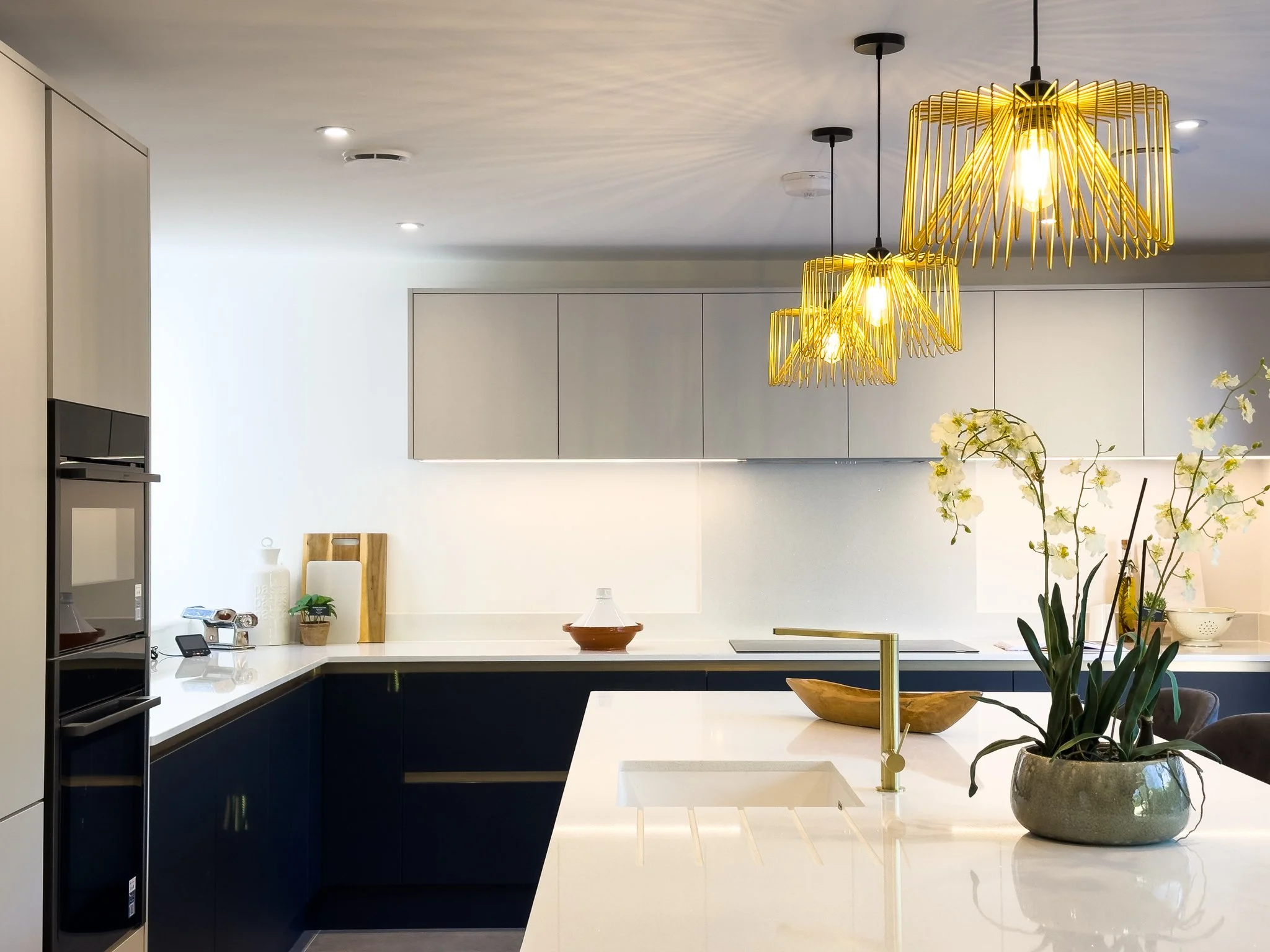 Modern kitchen with white countertops, navy blue lower cabinets, and white upper cabinets. Three gold pendant lights hanging above the island, decorated with a potted orchid plant and a wooden bowl. Recessed ceiling lights and a built-in oven are als