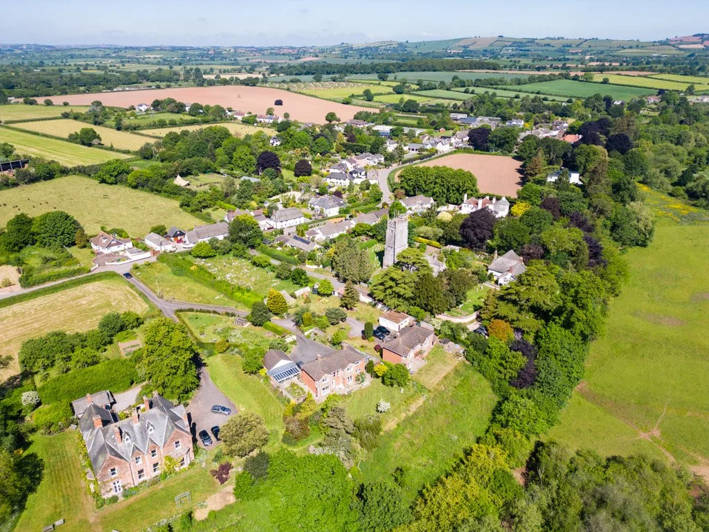 Aerial view of a small village with houses, a church, and surrounding lush green fields and trees.