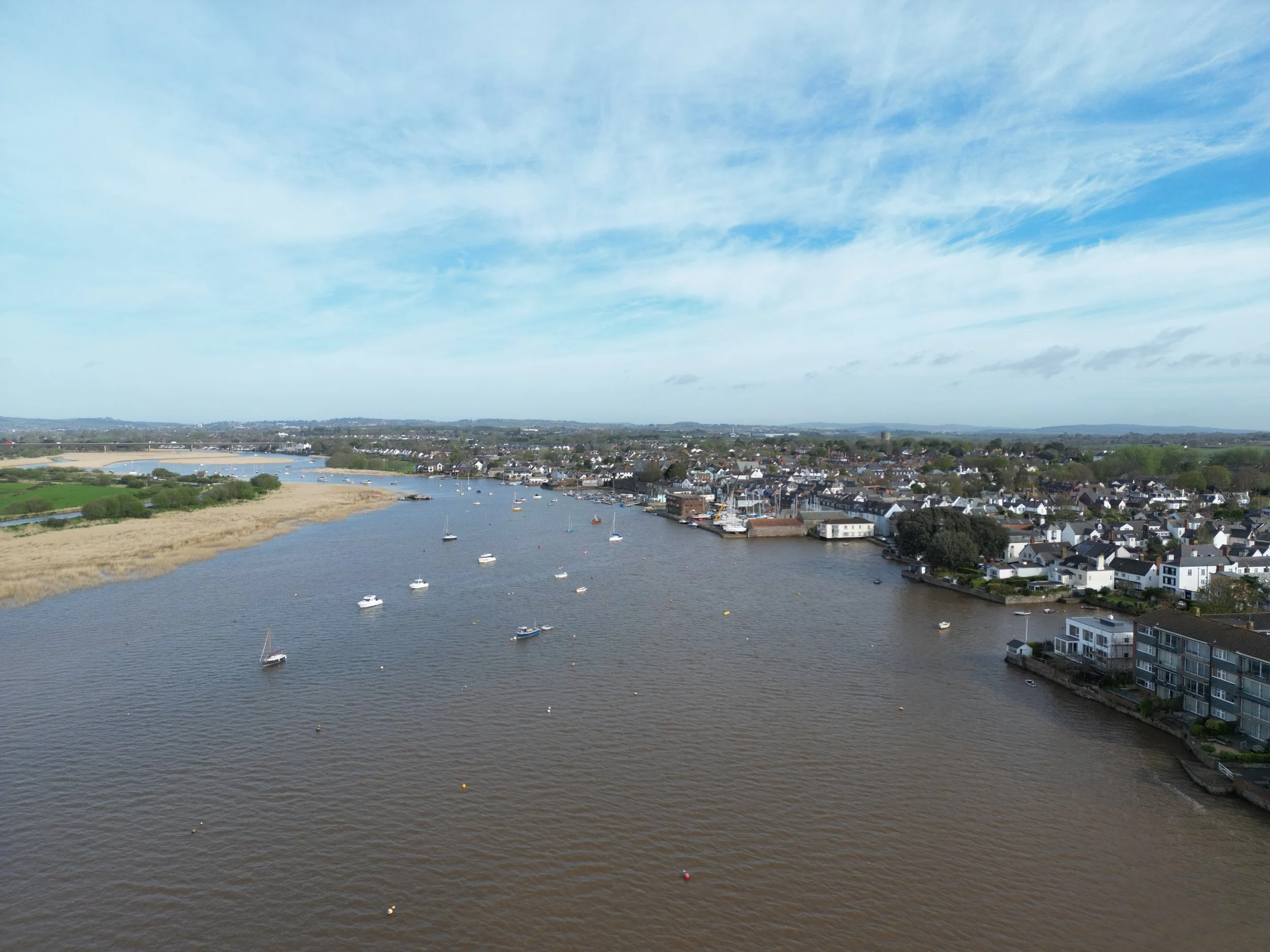 Aerial view of a river with multiple sailboats and a town with white houses along the shoreline, under a partly cloudy sky.